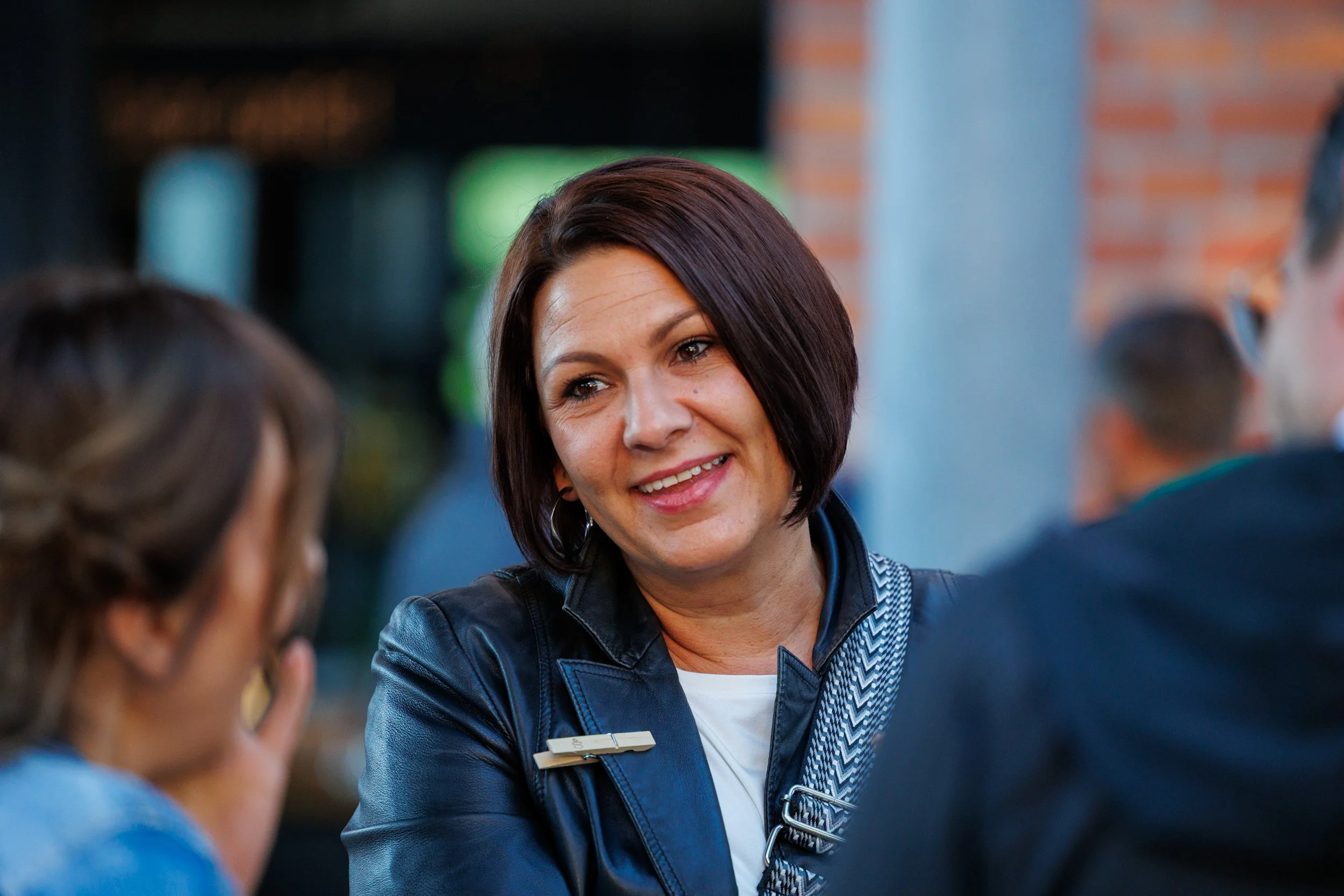 Eine fröhliche Frau im Gespräch mit zwei anderen Personen in einem öffentlichen Umfeld, vermutlich bei einer Veranstaltung oder einem Treffen. Eventfotografie