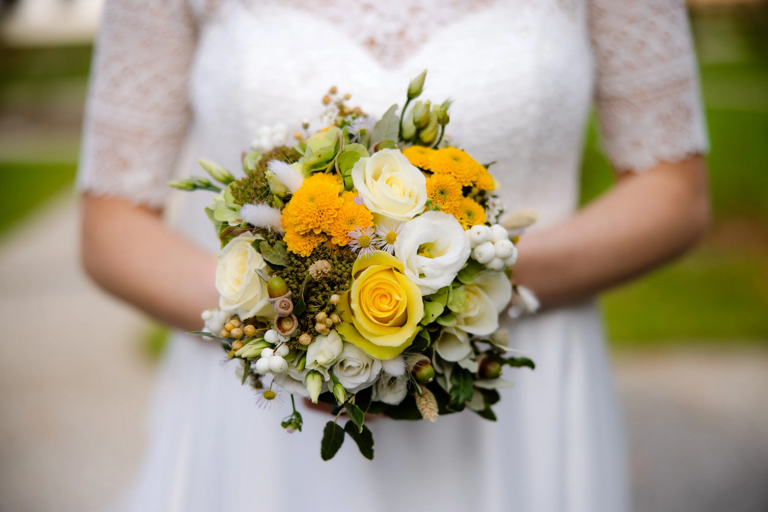 Frau in weißem Brautkleid hält einen Brautstrauß aus weißen und gelben Blumen, umgeben von Grünblättern, im Freien. Hochzeitsfotografie