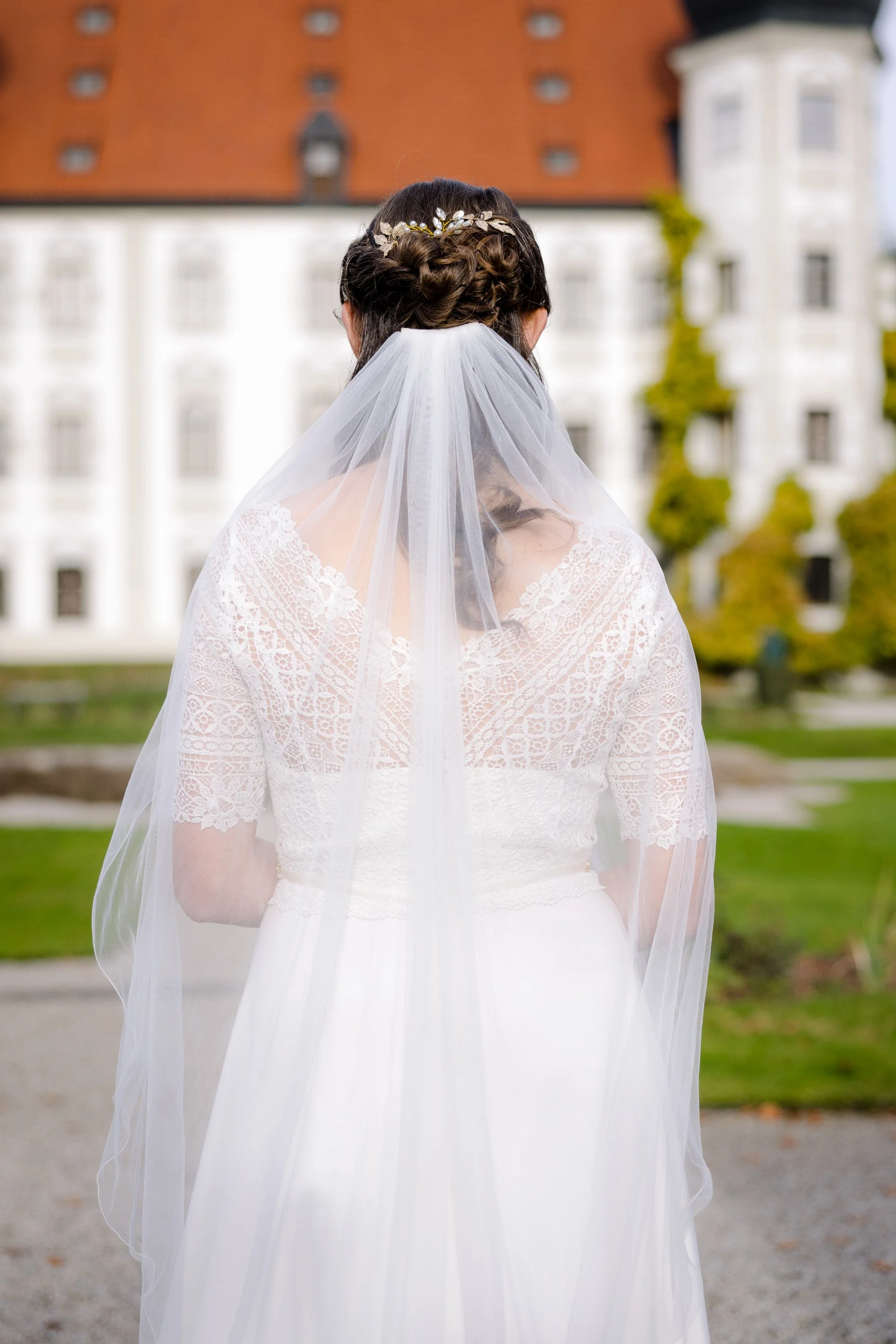 Braut in weißen Kleid mit Schleier und Haarschmuck, von hinten fotografiert, vor einem historischen Gebäude mit roten Dächern. Hochzeitsfotografie