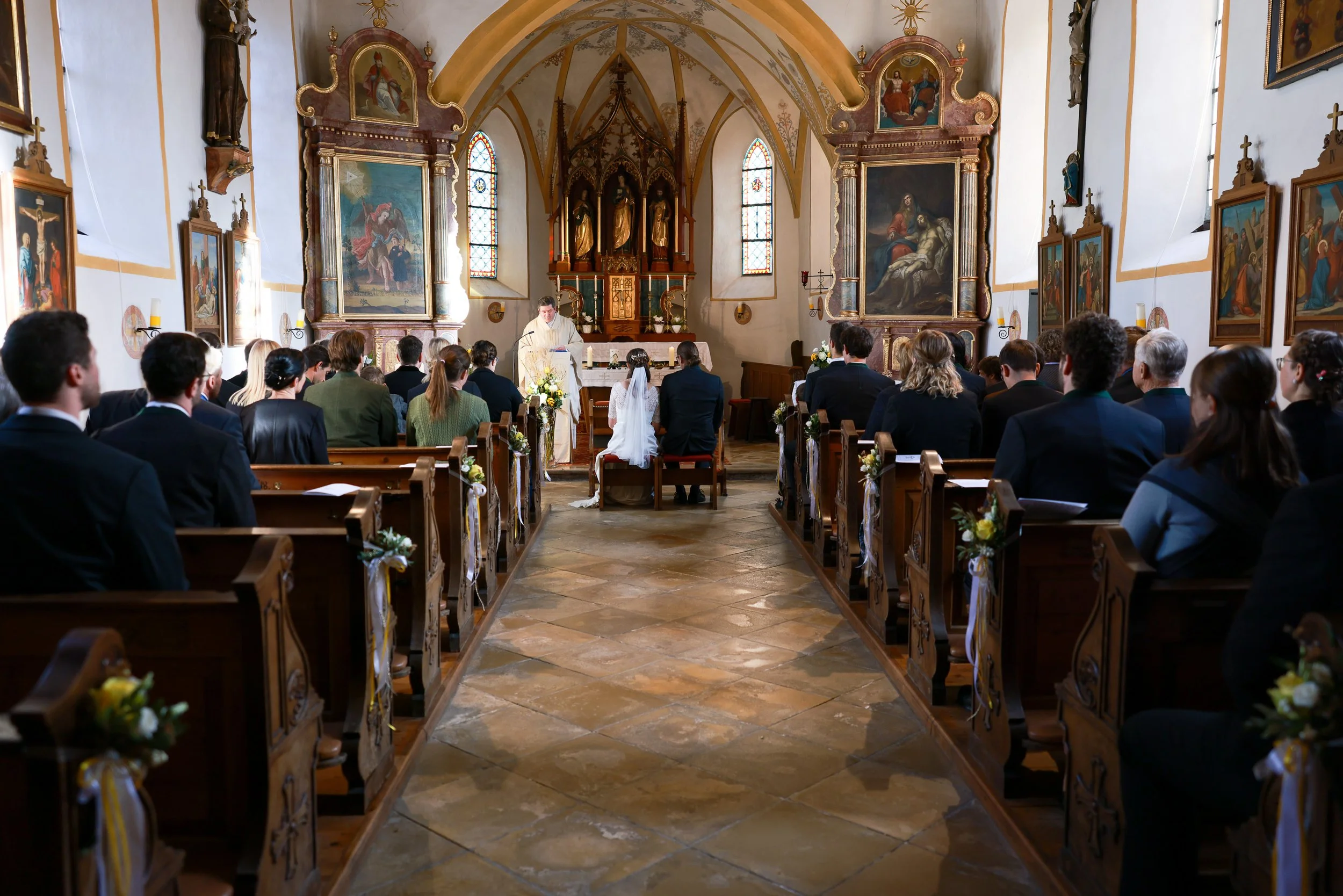 Hochzeit in einer Kirche mit einem Priester, Brautpaar sitzt vor dem Altar, viele Gäste sitzen in den Bänken, die Kirche ist mit religiösen Gemälden und Fenster mit buntem Glas dekoriert. Hochzeitsfotografie