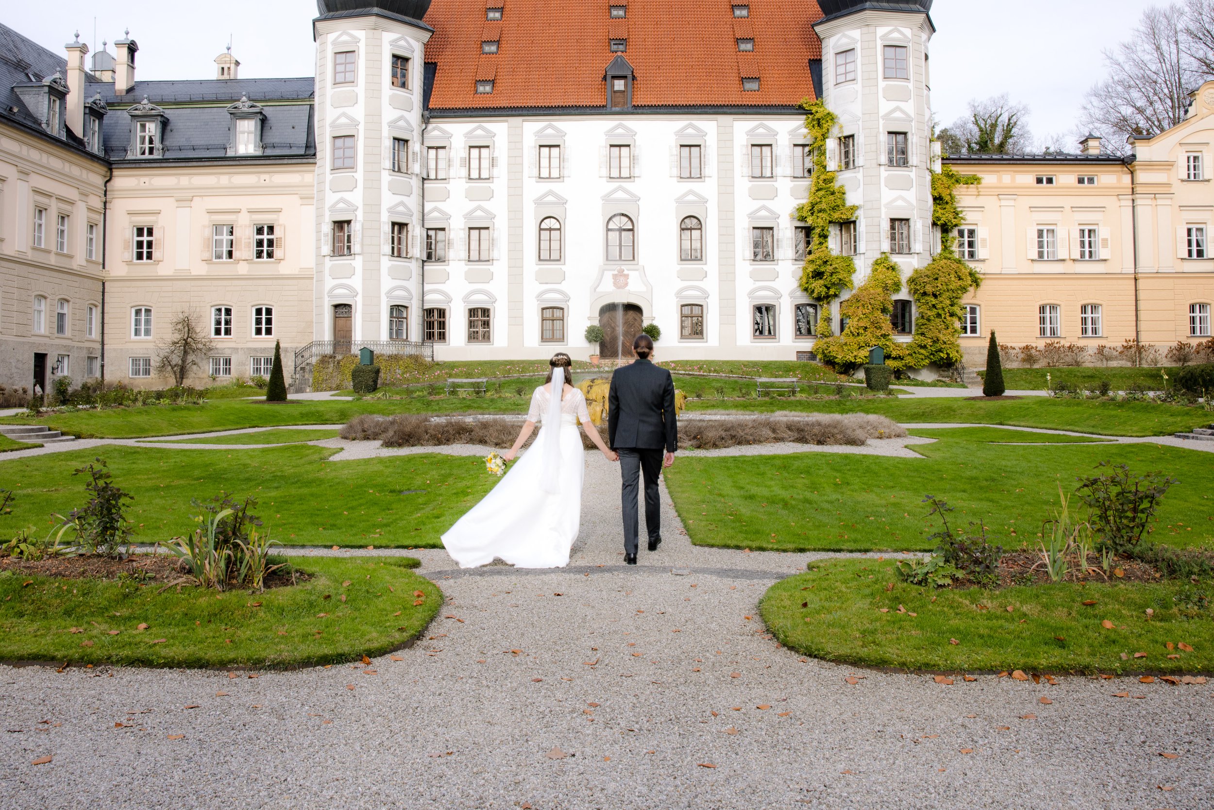 Ein brautpaar in Hochzeitskleidung, die Hand in Hand auf einen historischen Schlossgarten zugeht. Hochzeitsfotografie
