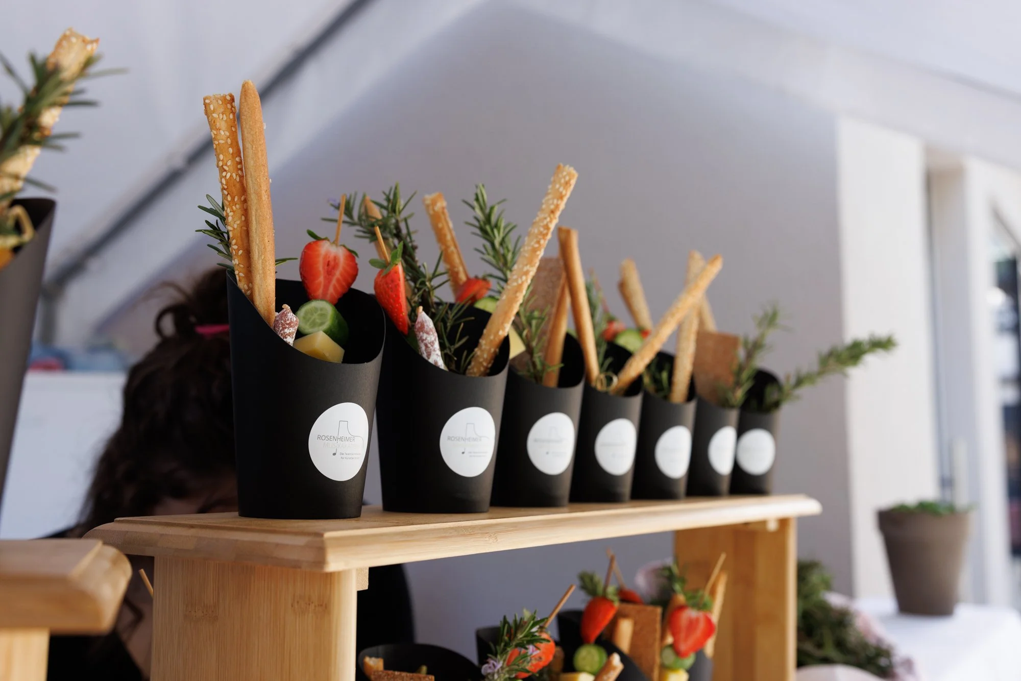 Schwarze Papiertöpfe mit Snacks, including Erdbeeren, Gurkenscheiben, Kräuter und Kekse, auf einem Holzregal. Eventfotografie