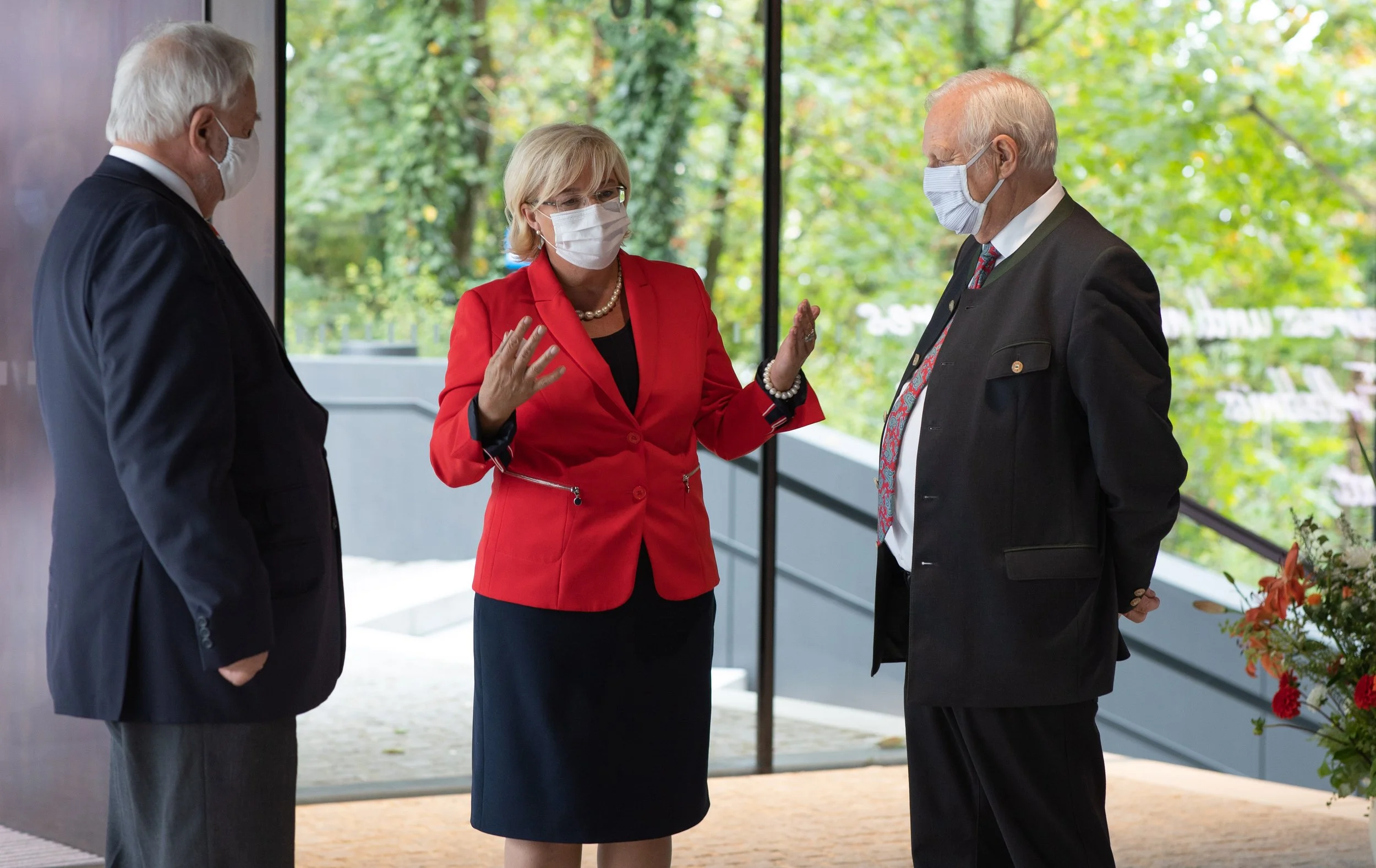 Drei Menschen im Gespräch, alle tragen Masken, im Hintergrund grüne Bäume sichtbar. Eventfotografie