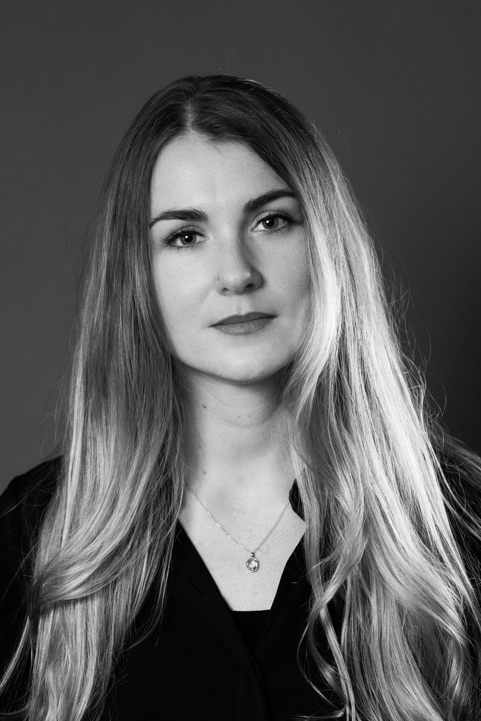 Schwarz-weißer Portrait einer jungen Frau mit langen, lockigen Haaren, die nach rechts fallen, und einem dunklen Hemd. Sie trägt eine Halskette mit einem Anhänger und hat einen kleinen Nasenpiercing. Porträt, Businessporträt