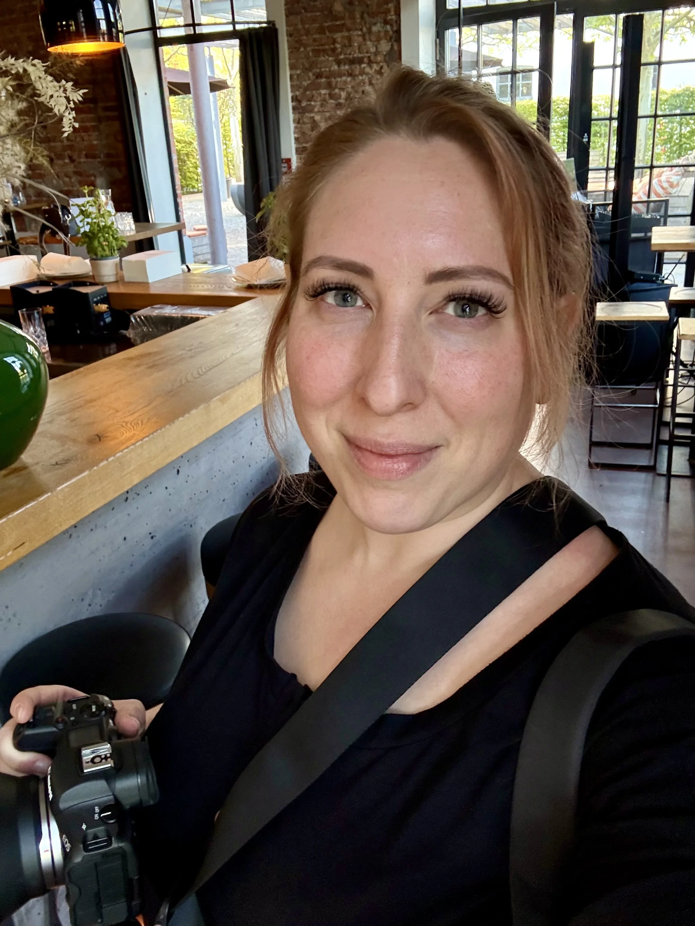 Eine junge Frau mit blonden Haaren und blau-grünen Augen, die eine Kamera in der Hand hält, posiert in einem modern eingerichteten Café. Im Hintergrund sind große Fenster, Holz- und Backsteinwände sowie Tische und Stühle sichtbar.