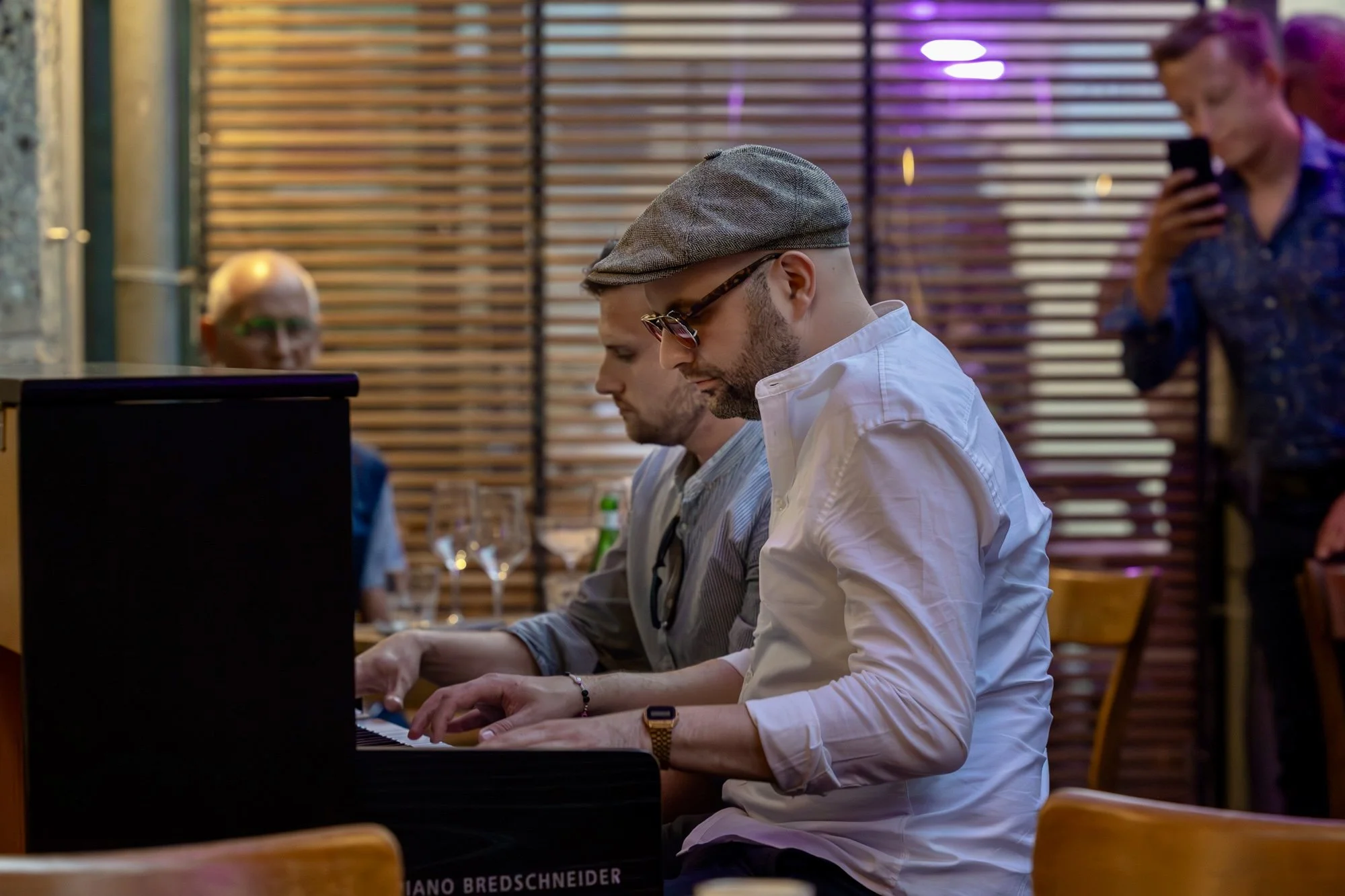 Zwei Männer spielen Klavier in einem Restaurant, eine Person im Hintergrund beobachtet, eine Person macht ein Foto, Wand mit Holzlamellen im Hintergrund. Eventfotografie