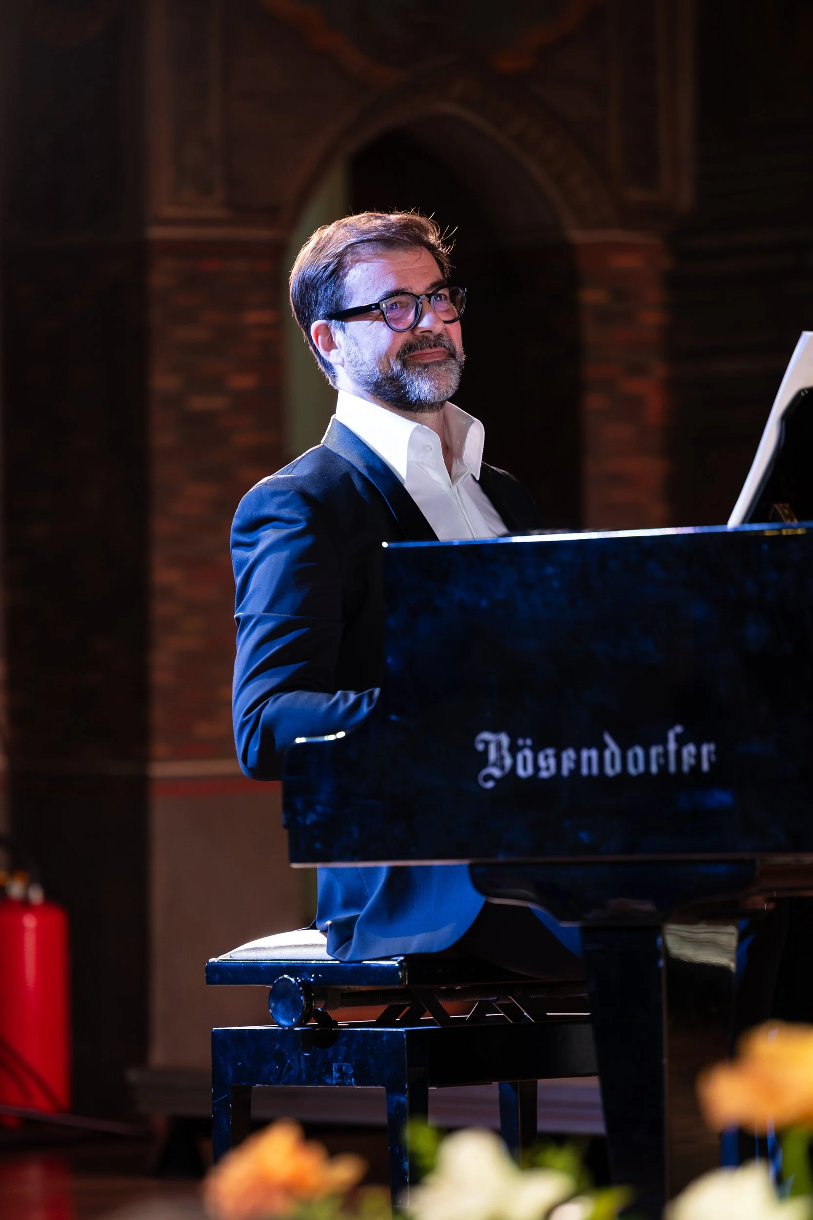 Ein Mann mit Brille und Bart spielt Klavier bei einer Veranstaltung in einem Raum mit dunklen Backsteinwänden. Eventfotografie
