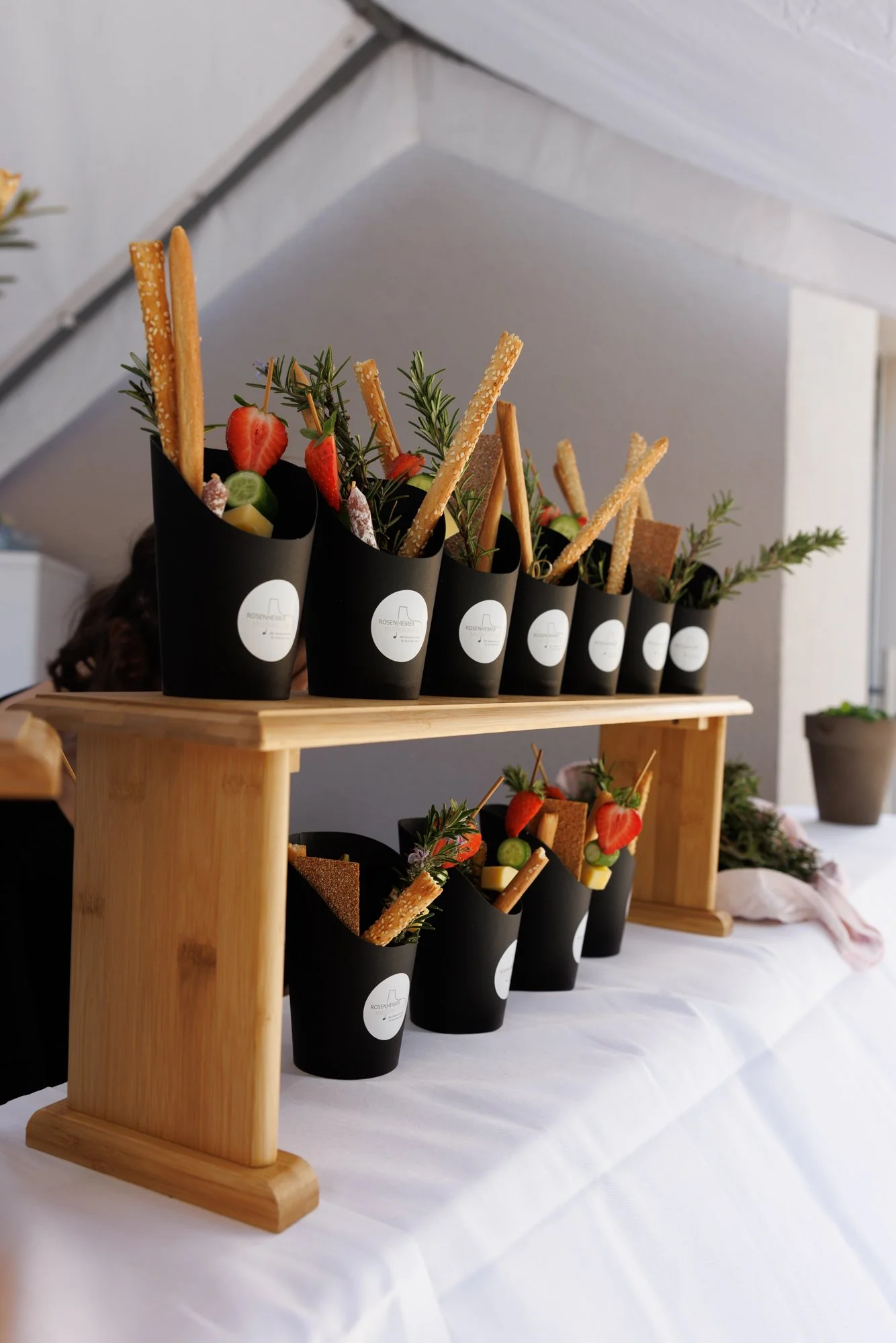 Mehrere kleine schwarze Töpfe mit Grüns, Erdbeeren, Gurken und Cracker, auf einem zweistöckigen Holzgestell auf einer weißen Tischdecke