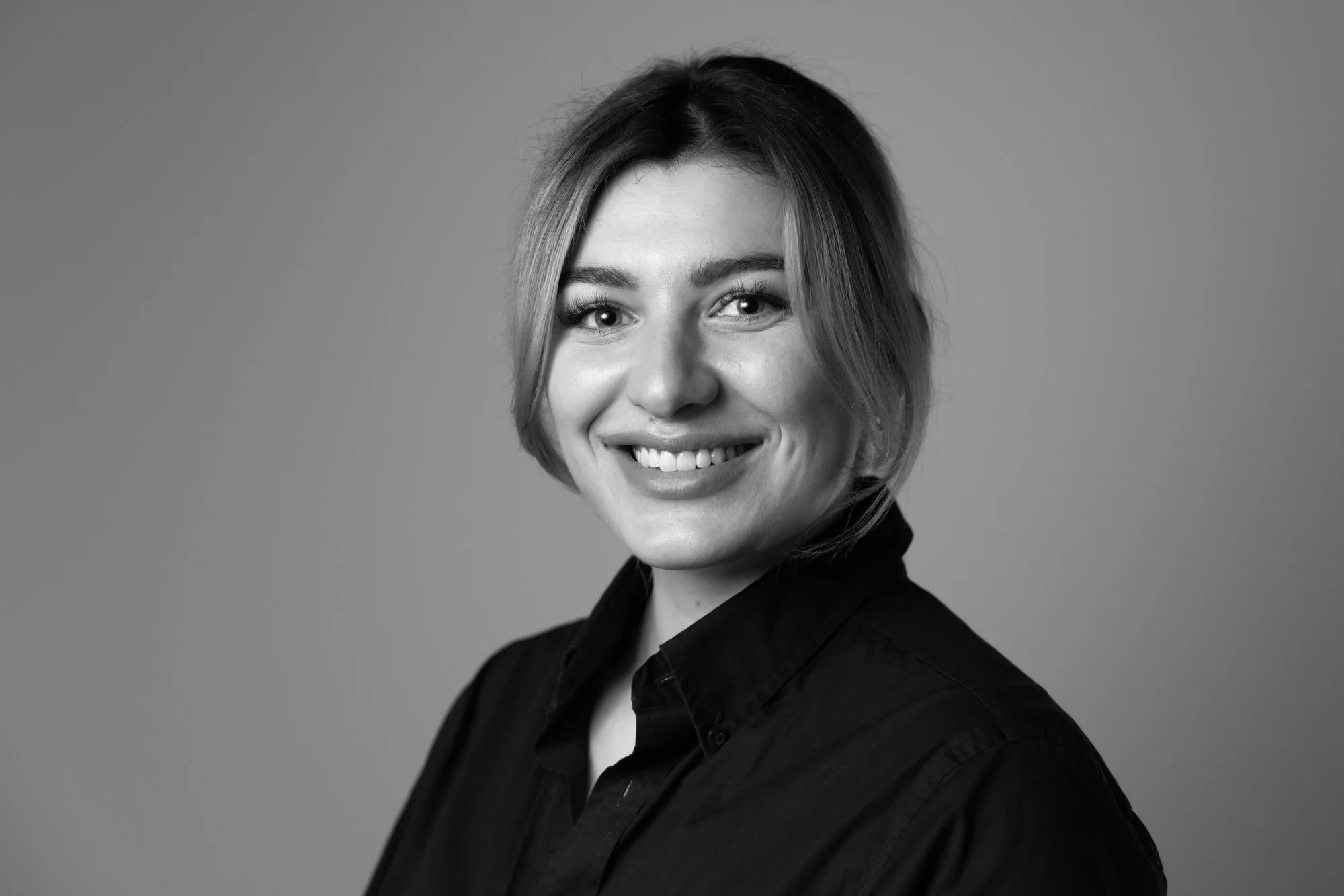 Schwarz-Weiß-Fotografie einer lächelnden jungen Frauenporträts, trägt dunkles Hemd, mit kurzem, gestylt Haar, vor neutralem Hintergrund. Porträt, Businessporträt