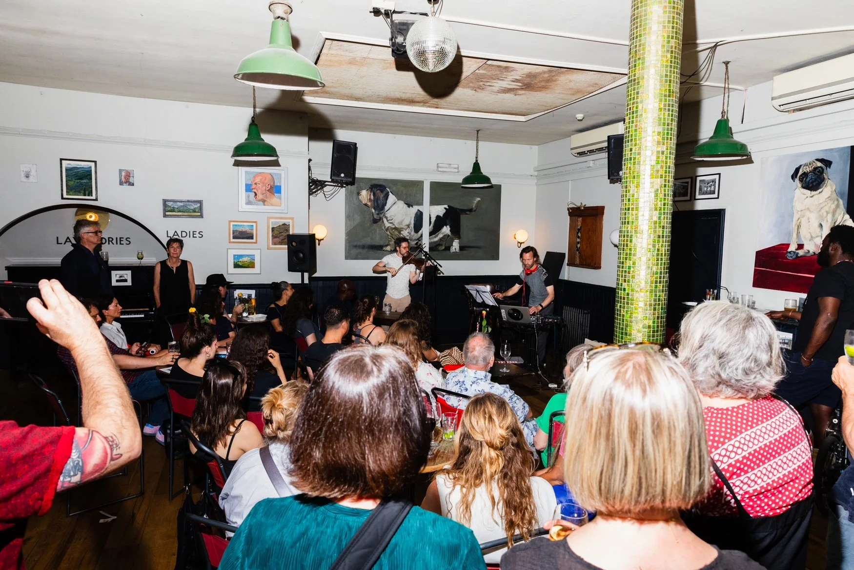 An indoor live music performance at a venue with an audience. Musicians on stage include a violinist and a DJ, with a singer in front. The walls are decorated with large dog paintings and framed photos. The audience is seated facing the performers.