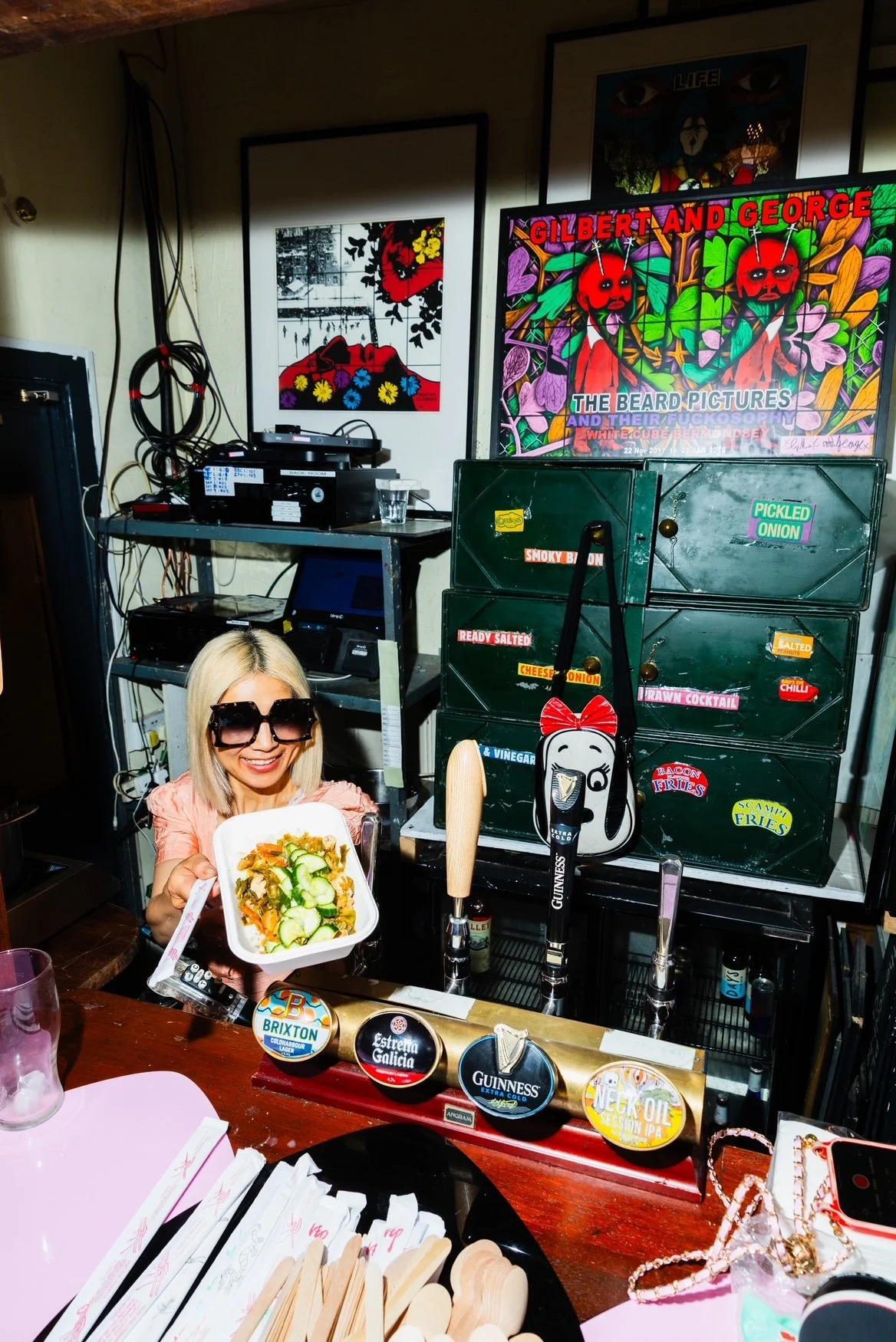 A woman with blonde hair wearing large sunglasses, smiling and holding a bowl of salad at a bar counter. Behind her, there are multiple tap handles for beer, a green storage container with colorful labels, television screens, and framed artwork on th