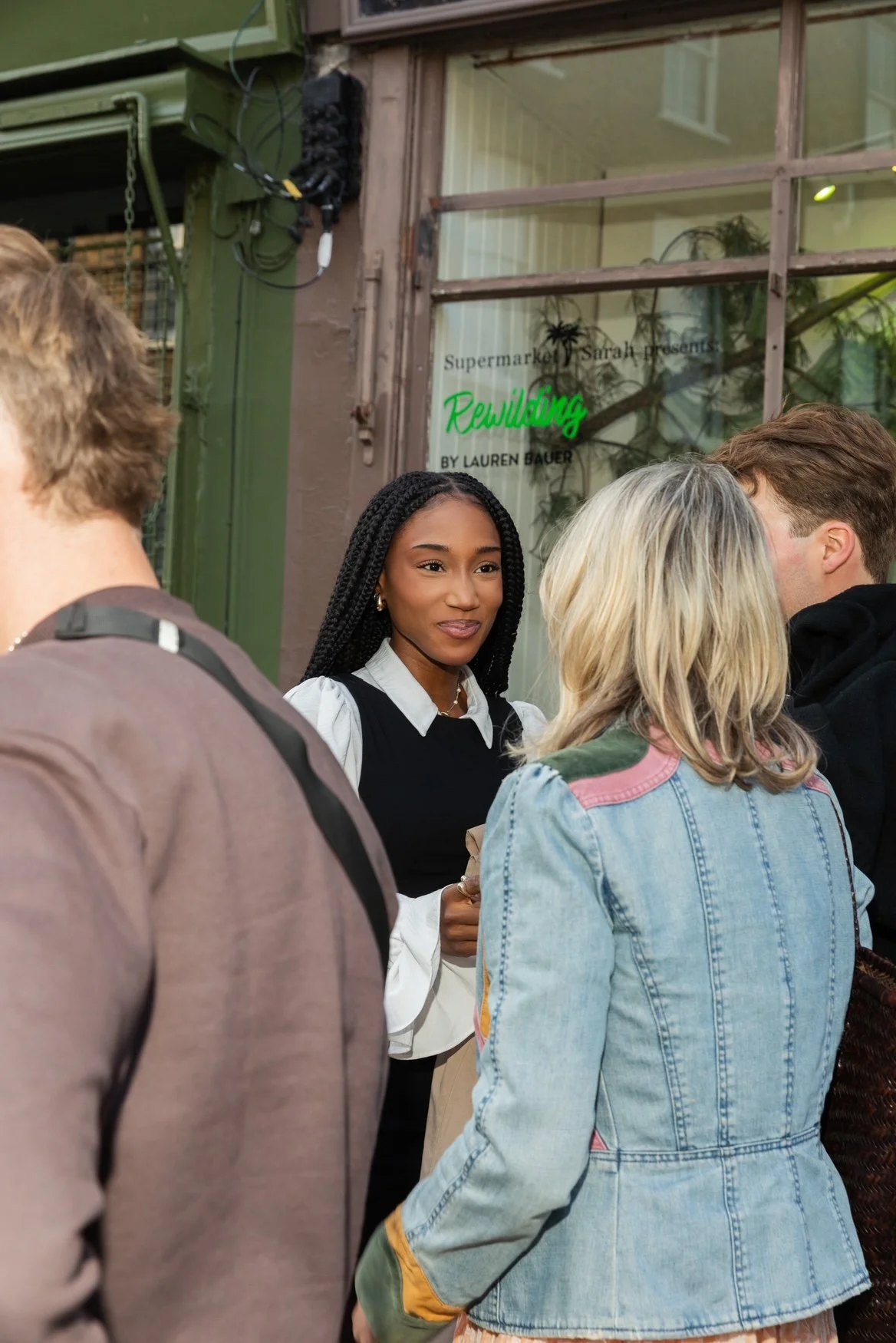 A group of people gathered outside a storefront with a sign that reads 'Supermarket Sarah presents Rewilding by Lauren Bauer'. One woman with braided hair, wearing a black vest over a white blouse, faces the camera while others have their backs turne