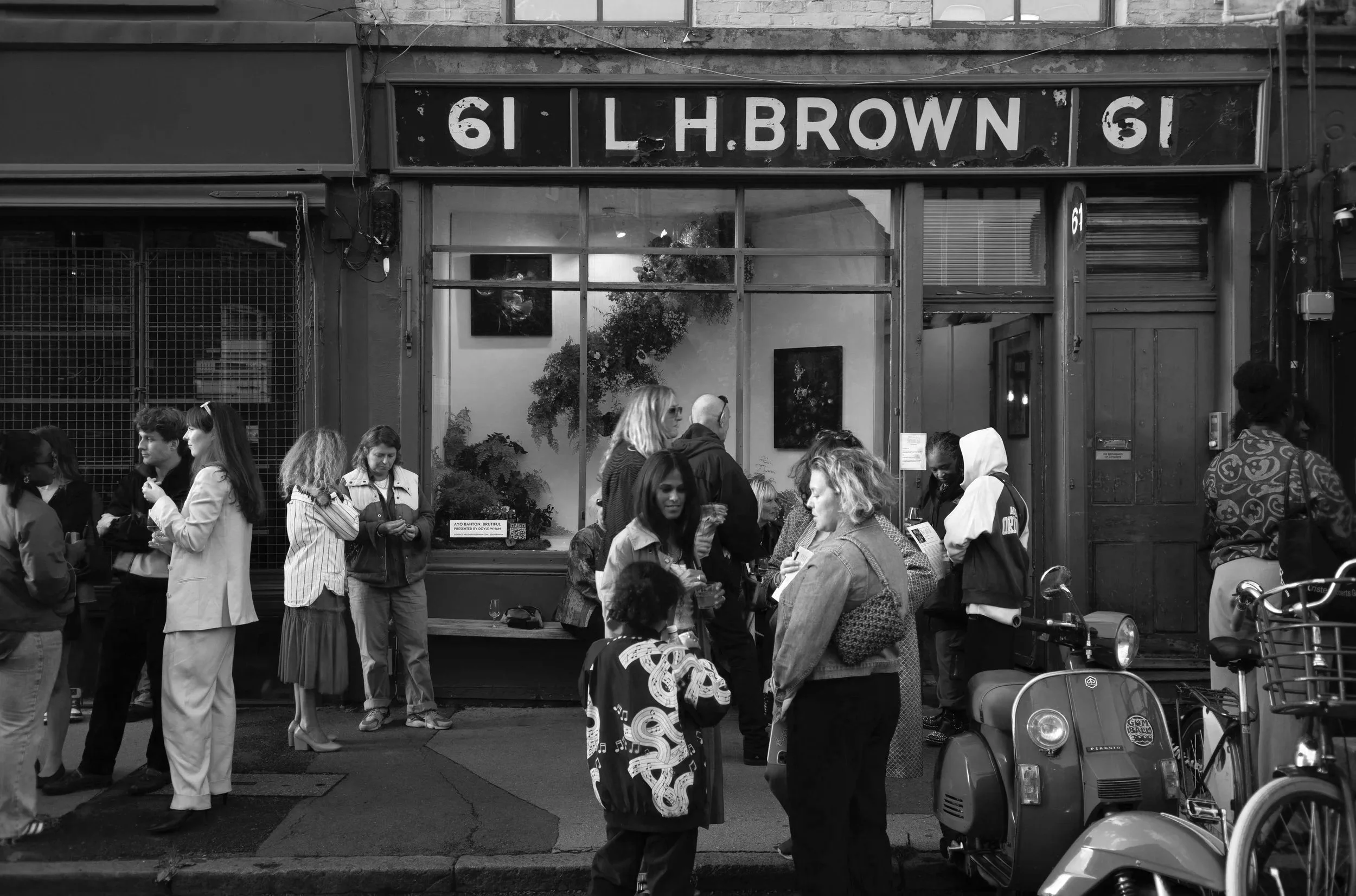 People gathered outside an art gallery with a sign display showing '61 L H BROWN 61'. The crowd includes women, men, and a child, some engaged in conversation and some looking at their phones. A scooter is parked in front of the gallery.