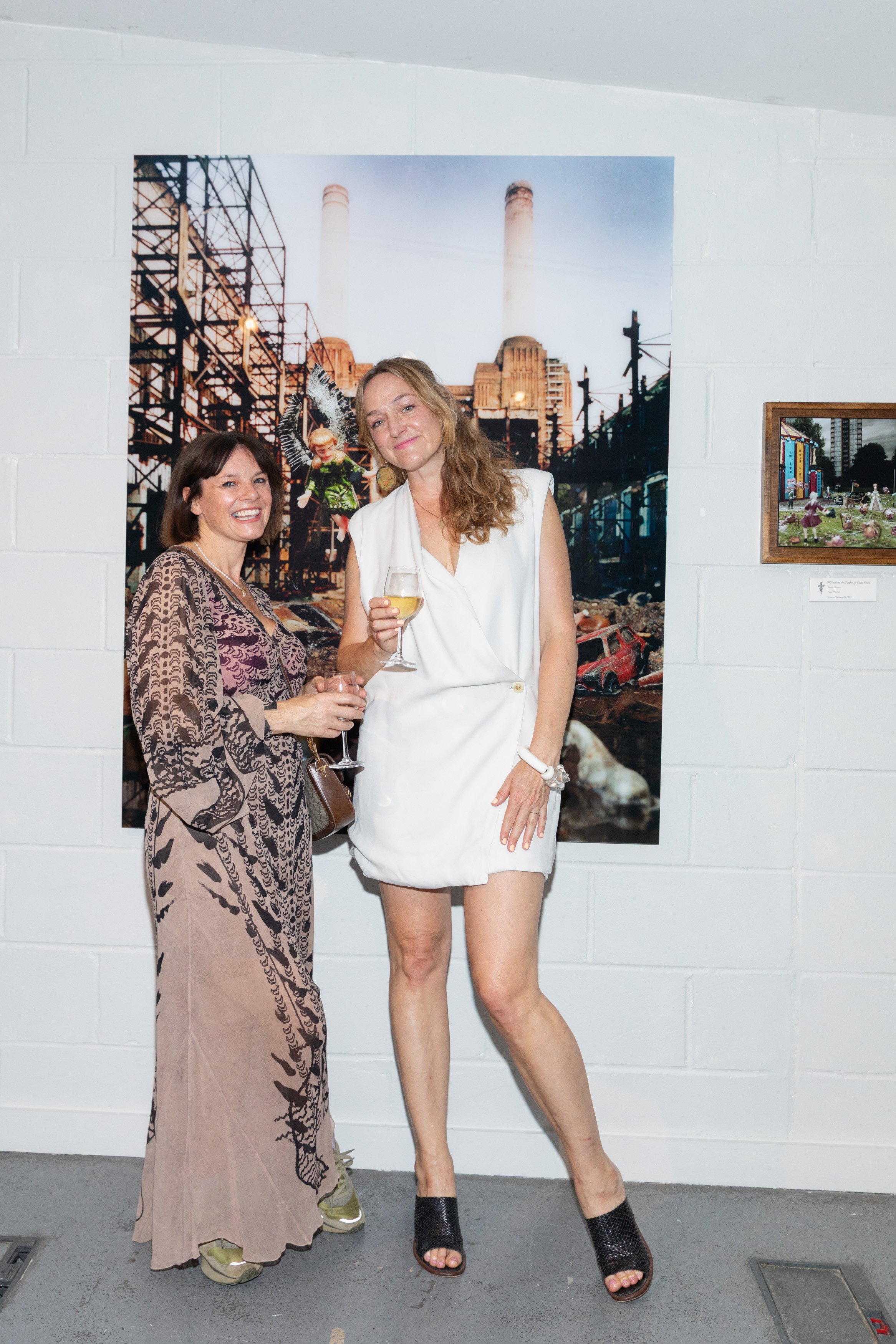 Two women in an art gallery standing in front of a large urban landscape artwork, holding glasses of white wine, smiling at the camera.