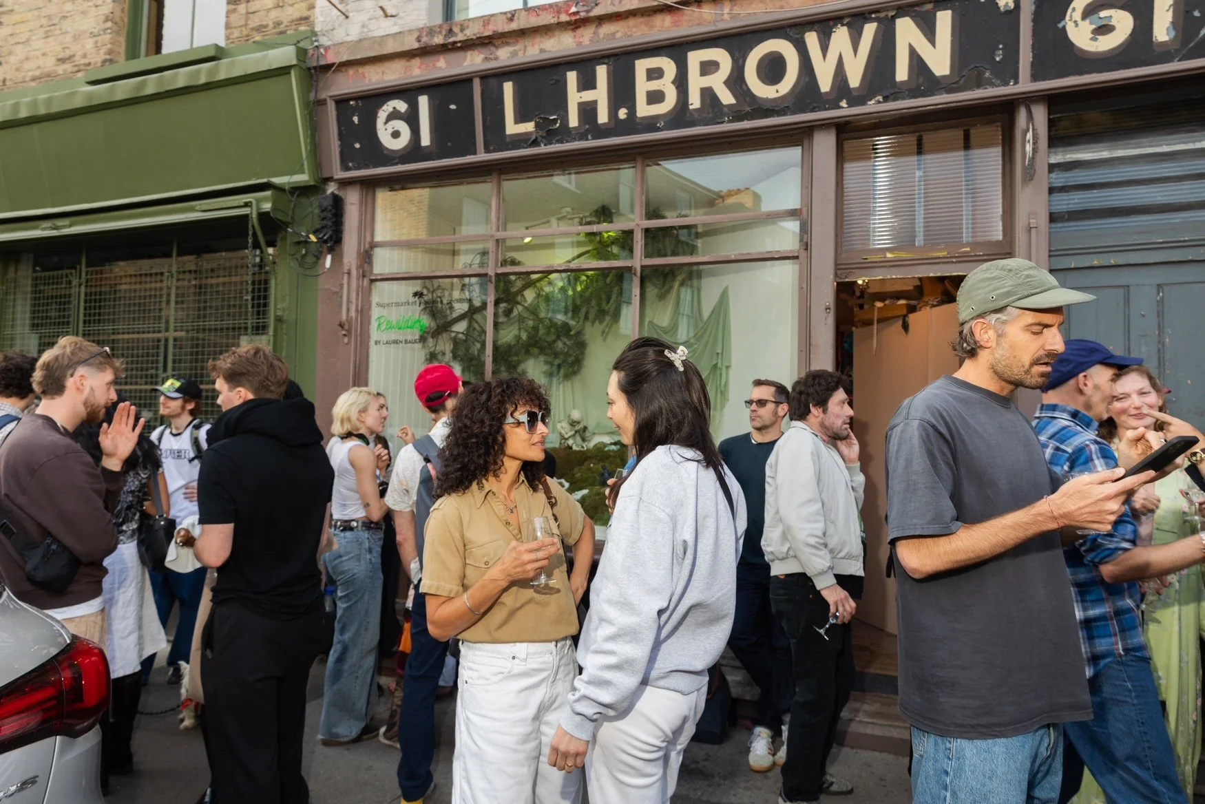 People gathered outside a building with a sign that reads '61 L. H. BROWN' and 'ST'. Some are talking, holding drinks, and using phones, while others wait or socialize.