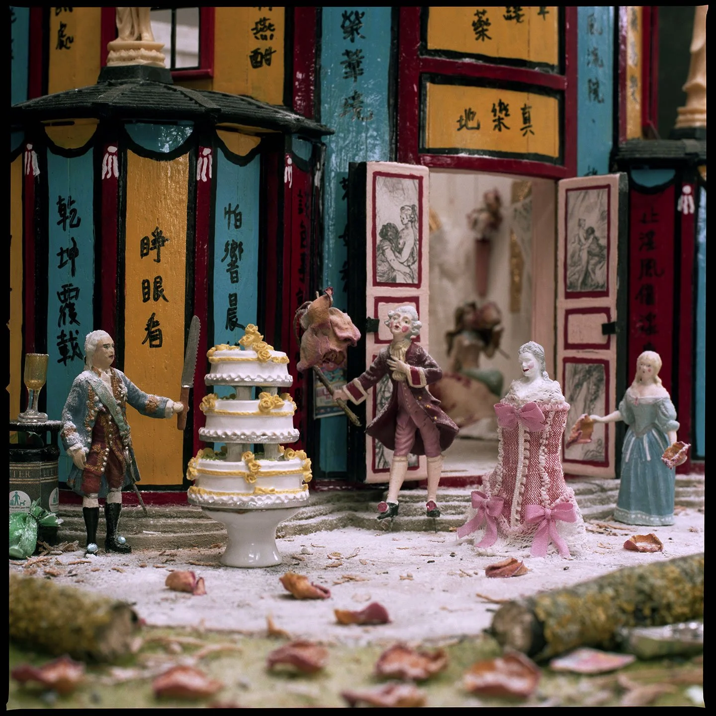 Miniature figurines of people in historical attire engaging in a celebration outside a colorful, Asian-themed building with Chinese characters, a tiered cake, and scattered leaves and debris on the ground.
