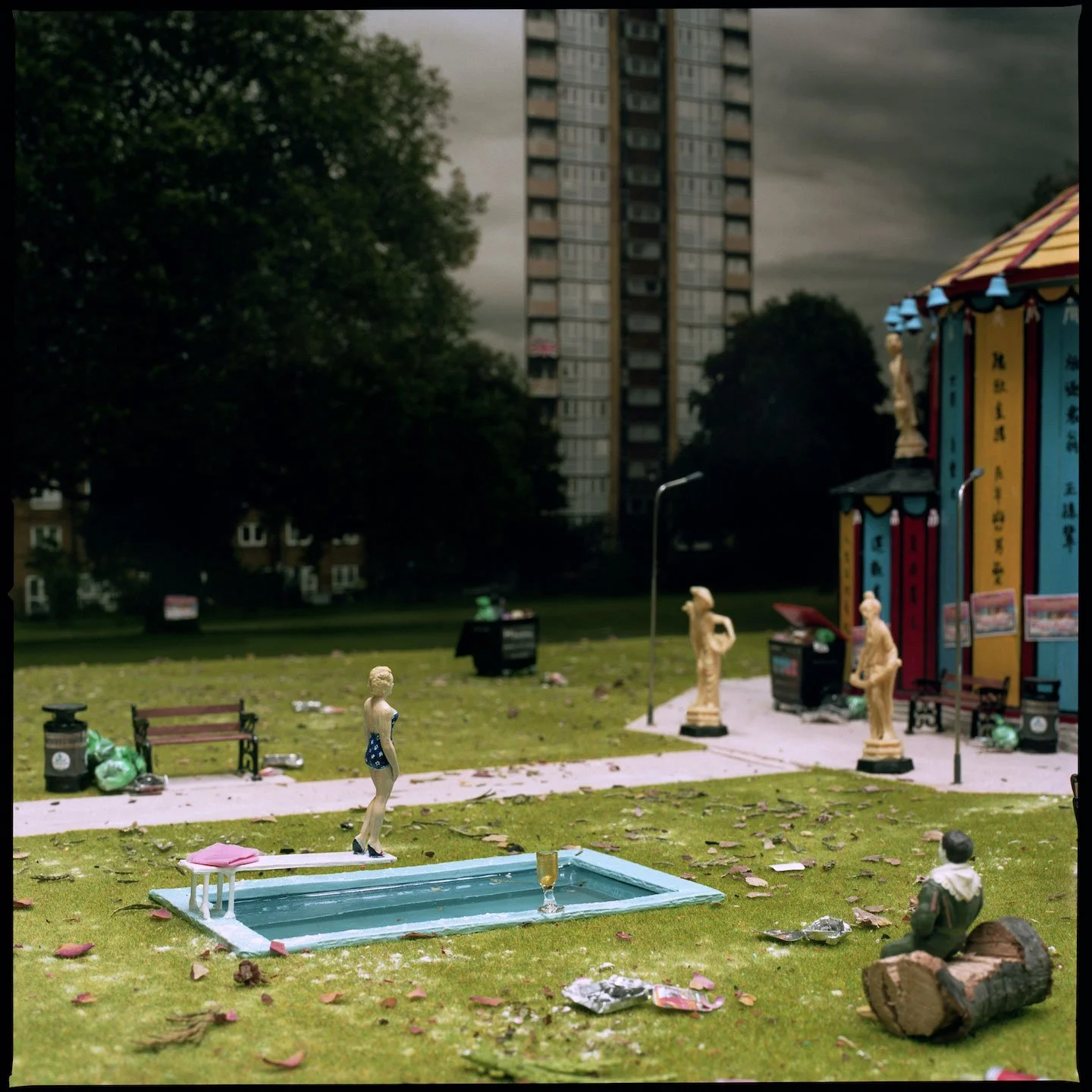 Miniature figurines of people gathered around a small pool in a park, with trash scattered on the grass and a colorful amusement ride in the background amid trees and high-rise buildings.