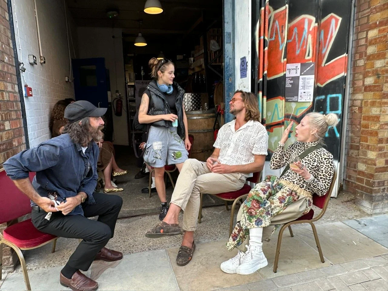 Four people having a conversation outside a building with graffiti on the door. One woman is standing, three people are sitting on maroon chairs; one man with long hair and glasses, a woman with white hair and animal print clothing, and another man w
