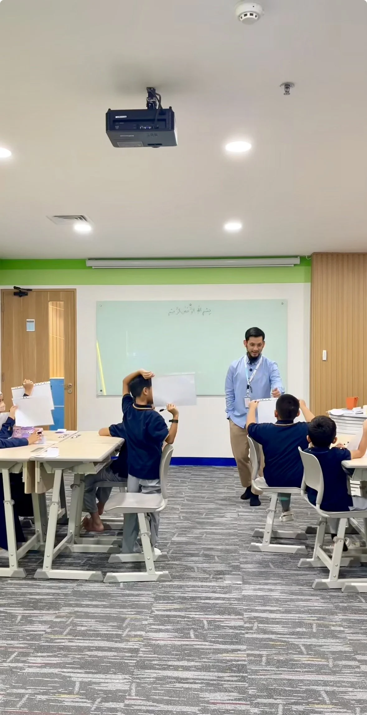 A classroom with students holding whiteboards and a teacher standing near a whiteboard, engaging with students during a lesson.
