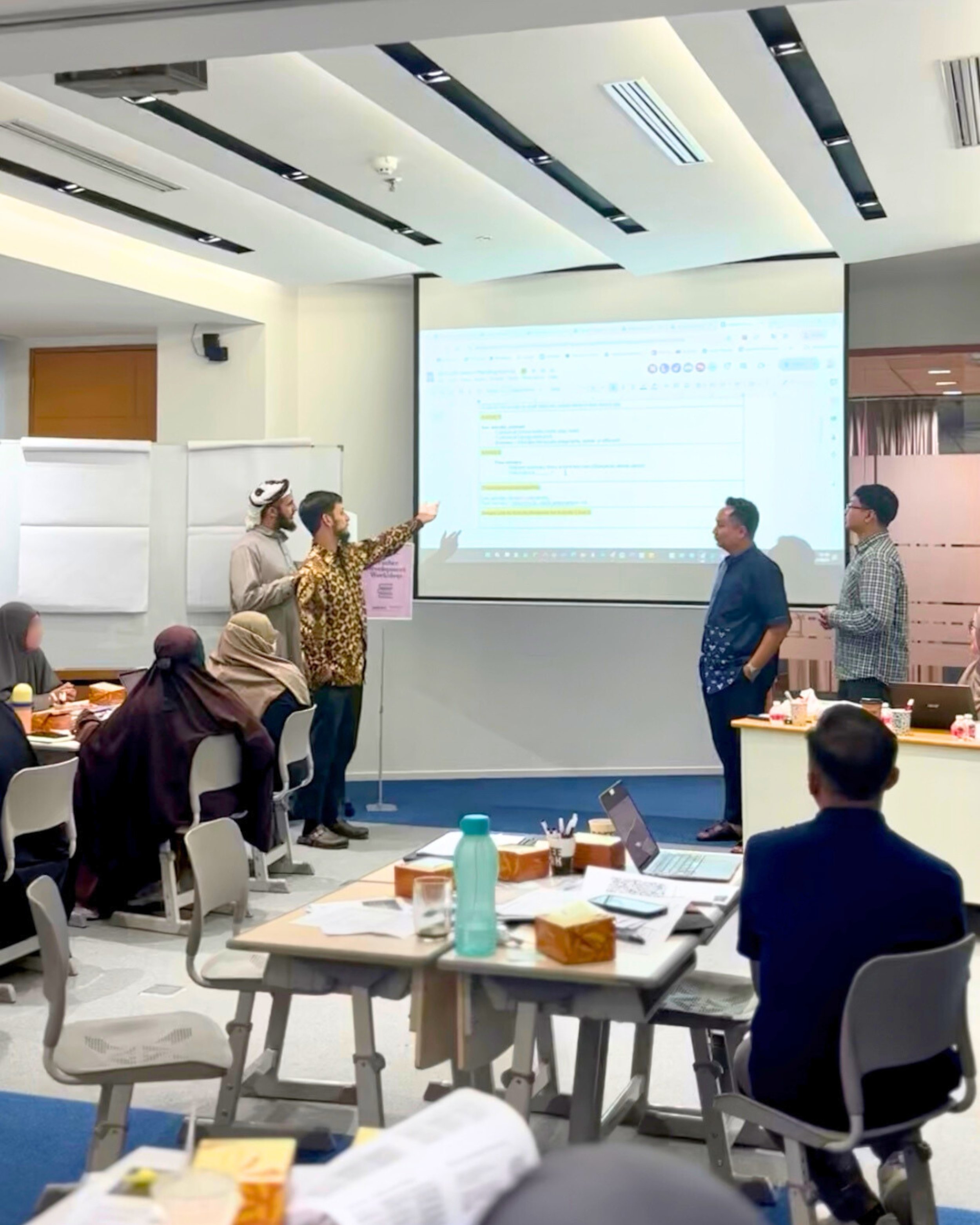 Business meeting or workshop in a conference room with people presenting and seated at tables, a large screen displaying a document or presentation, and laptops and refreshments on tables.