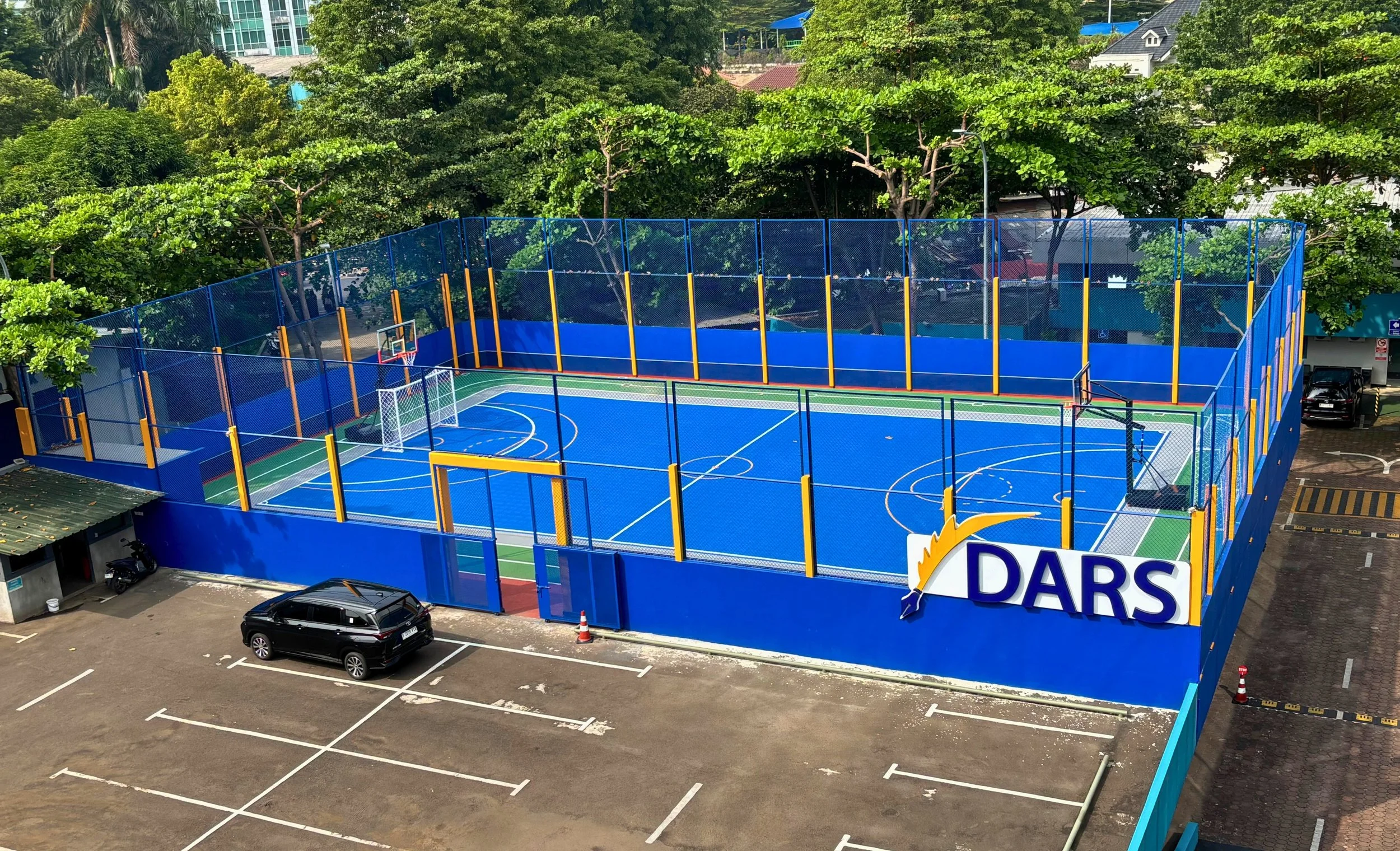 A colorful outdoor basketball and multi-sport court surrounded by a blue fence, with the DARS logo on the fence, located in an area with parking and green trees in the background.