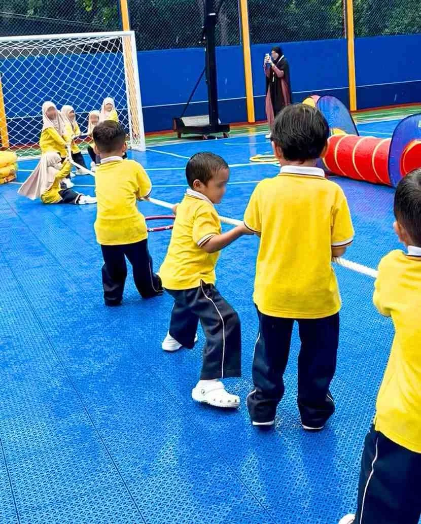 Children in yellow shirts playing tug-of-war on a blue indoor sports court, with a goal post and a person in the background taking a photo.
