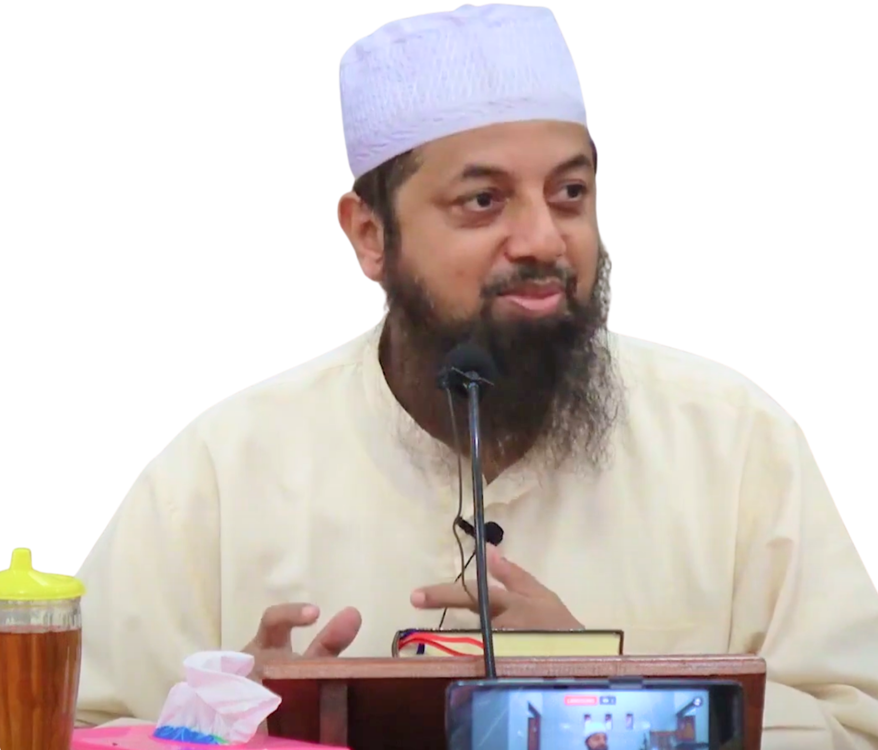 Man with a beard and white prayer cap speaking at a podium with a microphone, in front of a camera recording him.