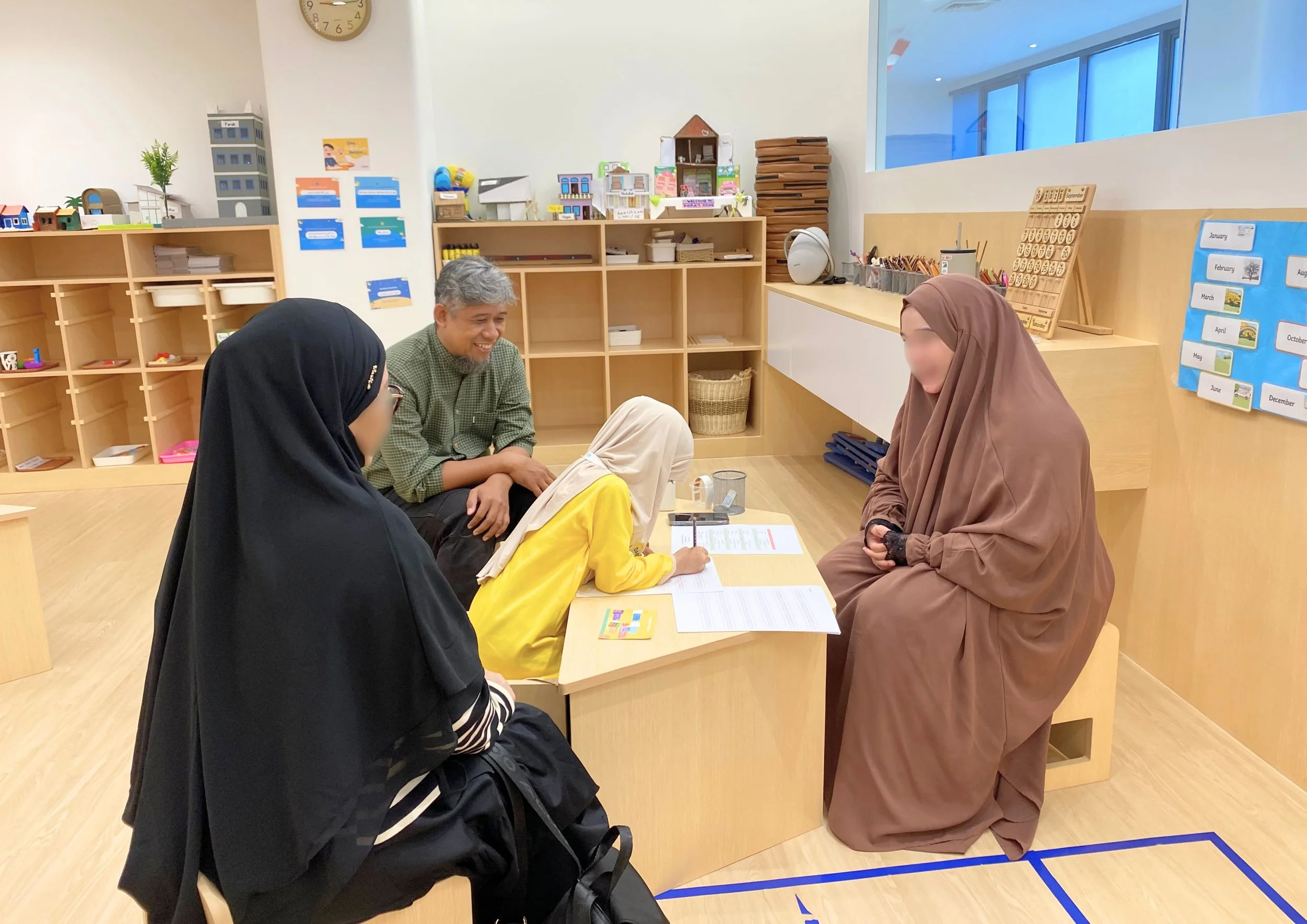 A group of four people, including children and adults, sit together in a classroom, engaging in a learning activity with papers and writing materials on the table. The background shows shelves with educational supplies and colorful learning materials.