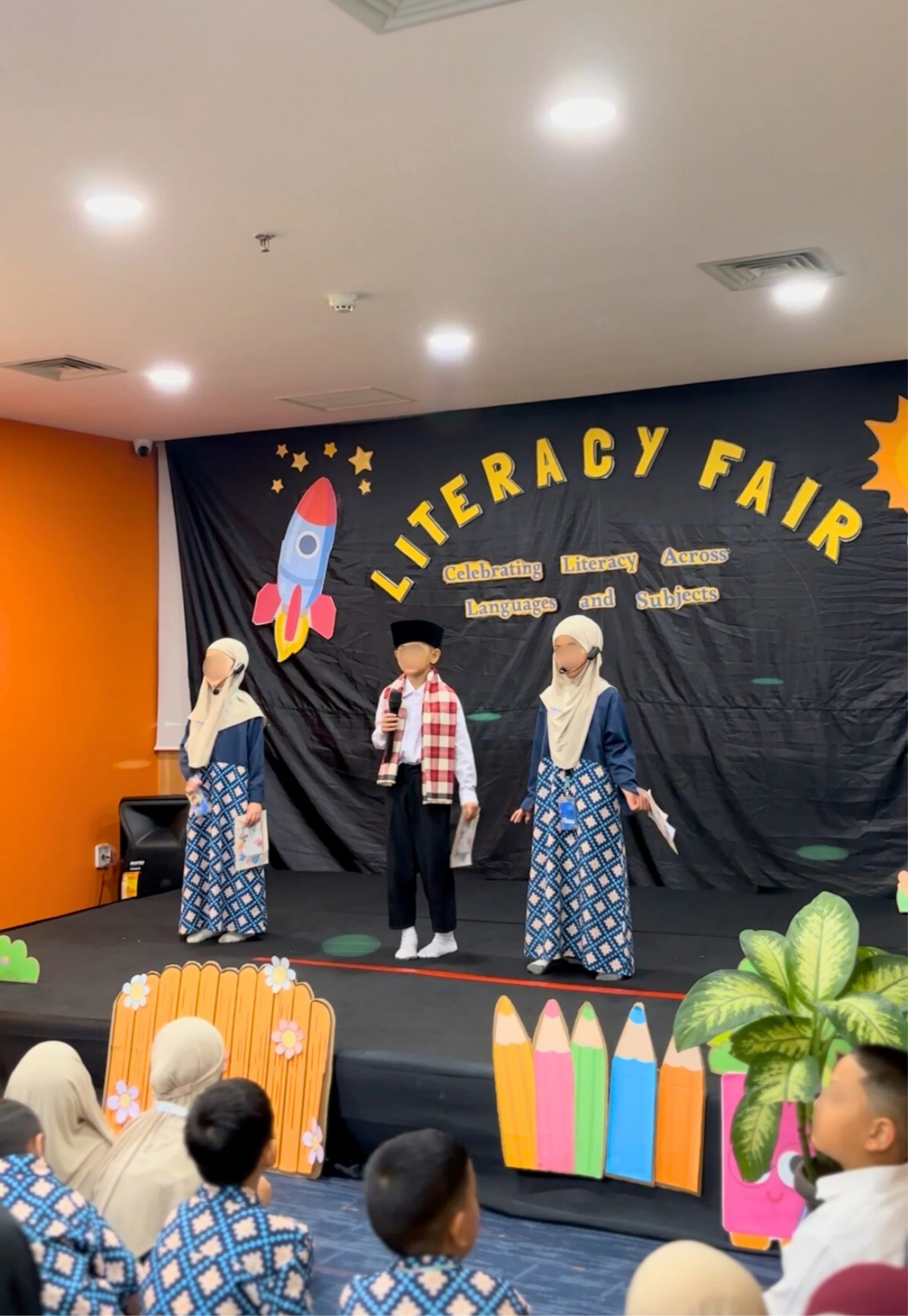 Children dressed in traditional attire and hijabs are performing on stage at a literacy fair, with a backdrop that reads 'Literacy Fair: Celebrating Literacy Across Languages and Subjects,' decorated with a rocket, stars, colorful pencils, and plants.