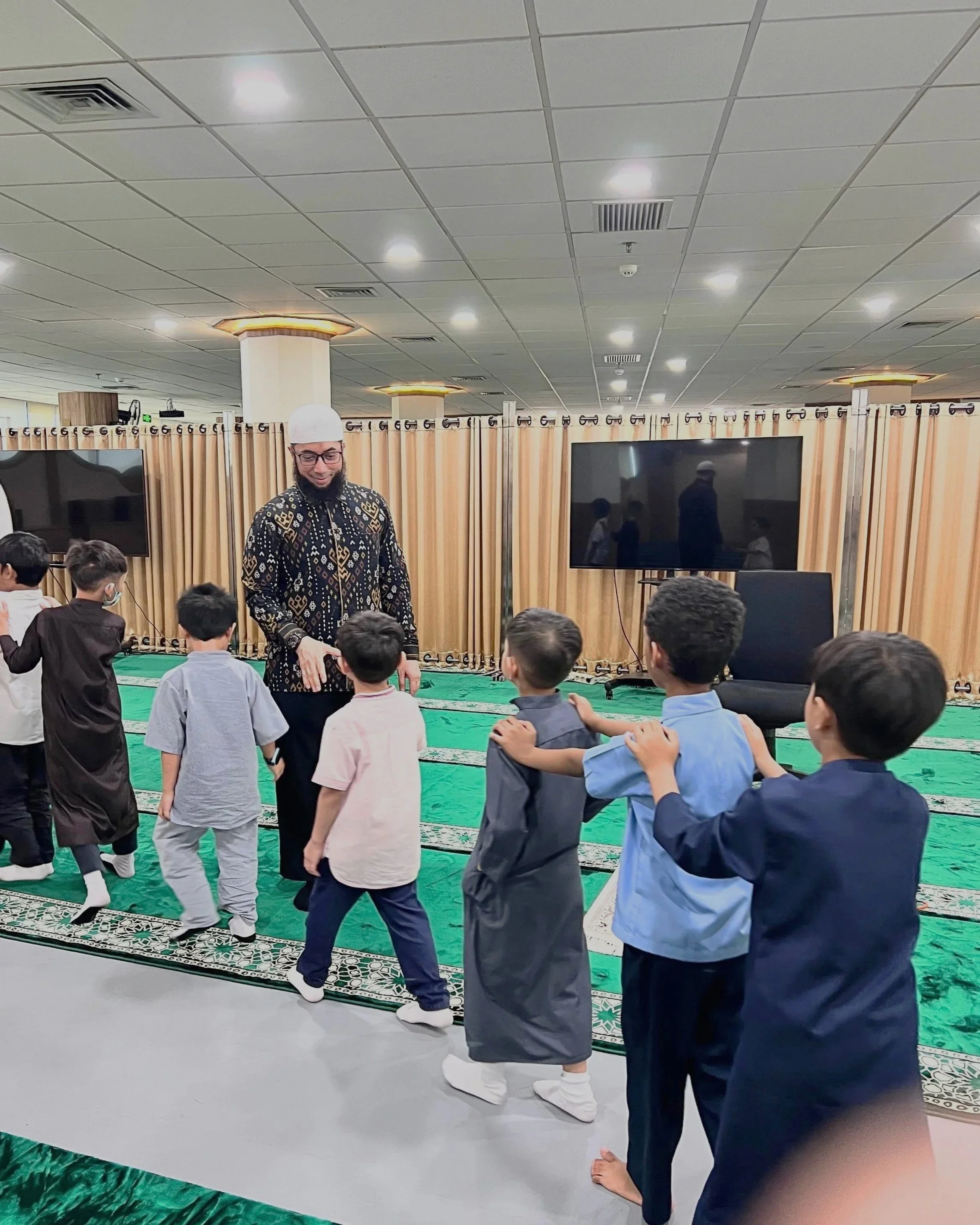 A man leading children in a group activity inside a room with green carpet, beige curtains, and two large flat-screen televisions.
