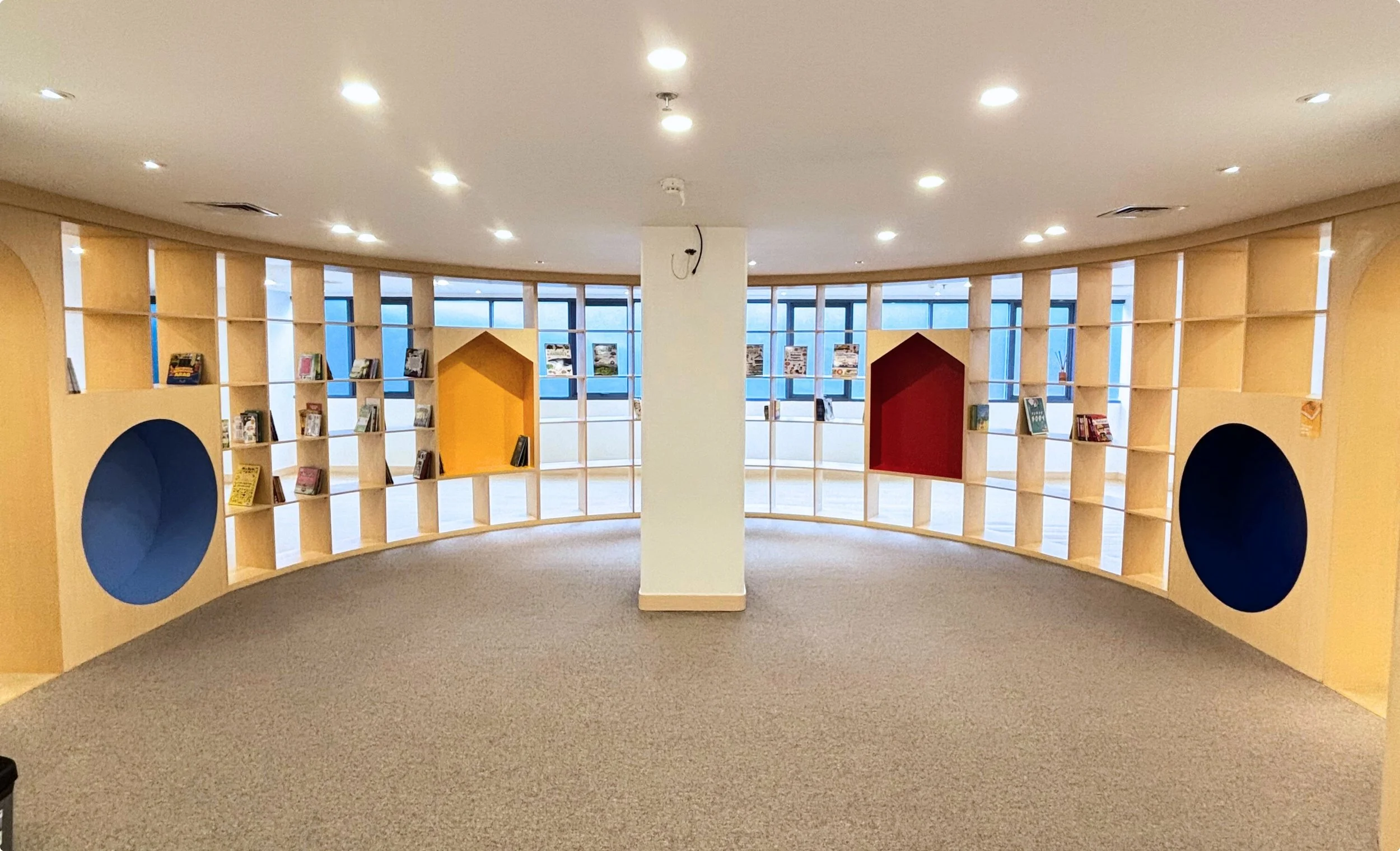  curved wooden bookshelf with circular and house-shaped openings filled with books, set in a spacious room with large windows and ceiling lights