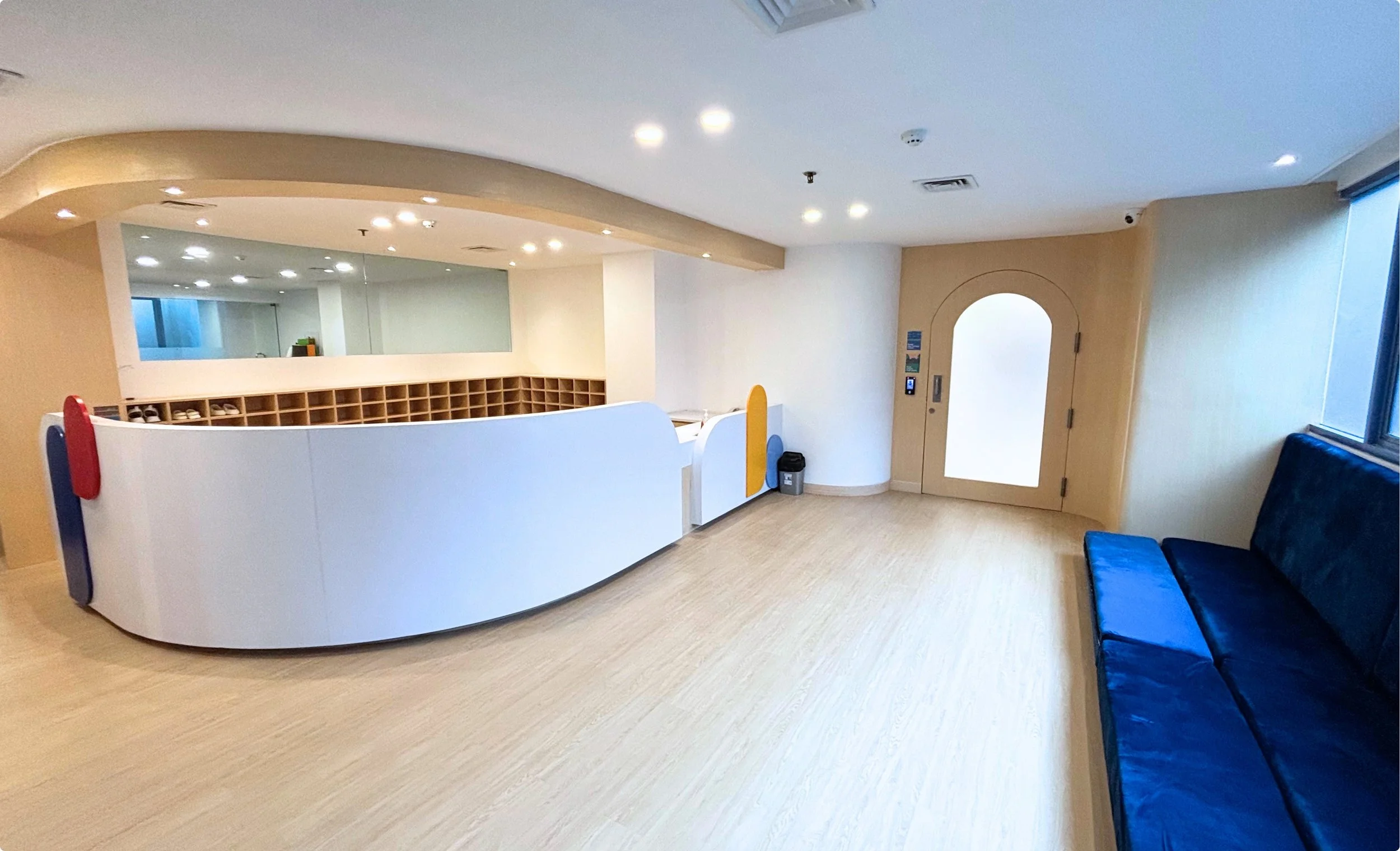 Empty modern reception area with a curved white desk, a blue bench, and a windowed door in a facility with wooden floors and ceiling lights.