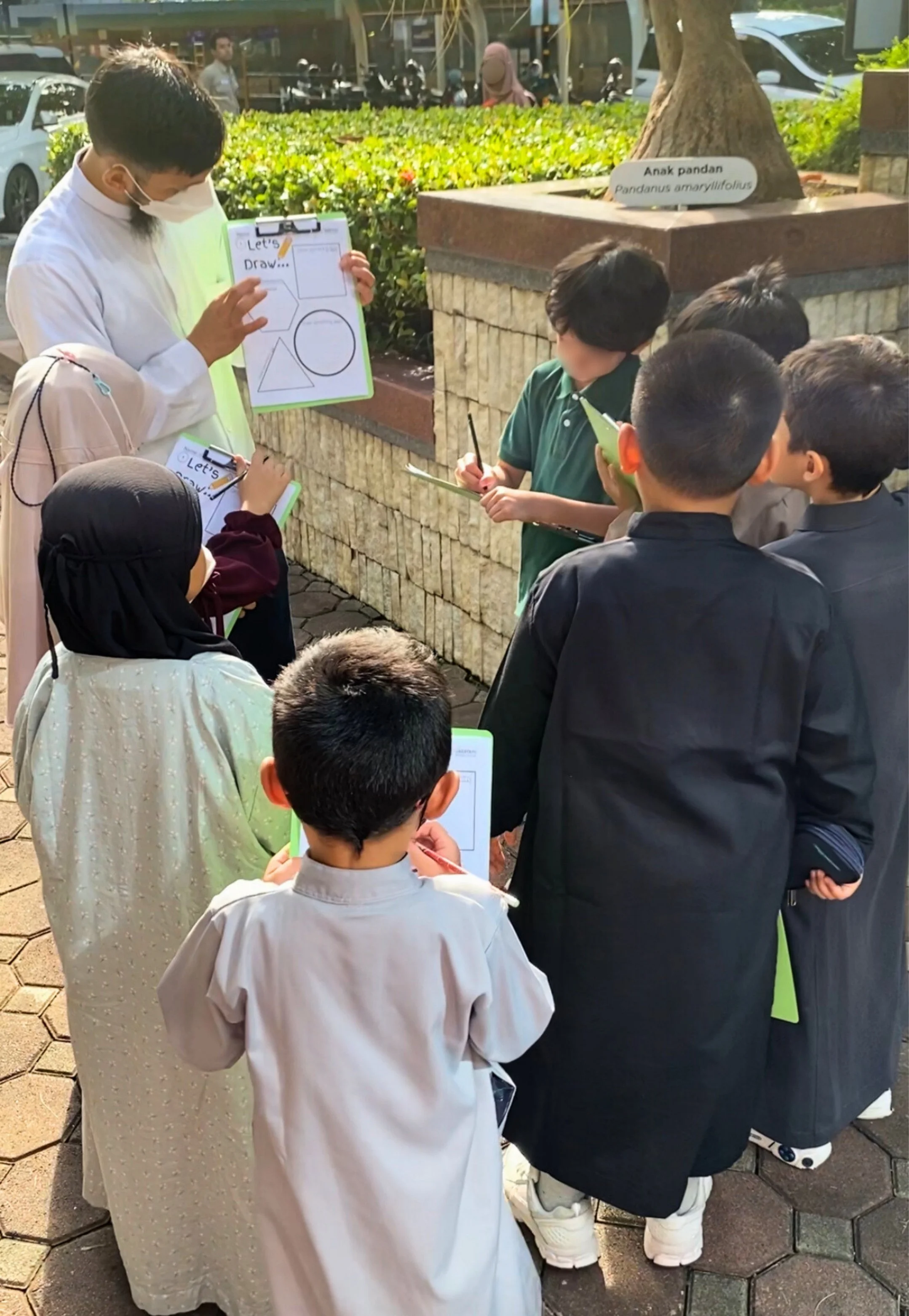 A group of children gathers outdoors around a person showing a drawing on a clipboard, with some children taking notes on tablets. A sign on a nearby brick planter identifies a tree as Pandanus amaryllifolius.
