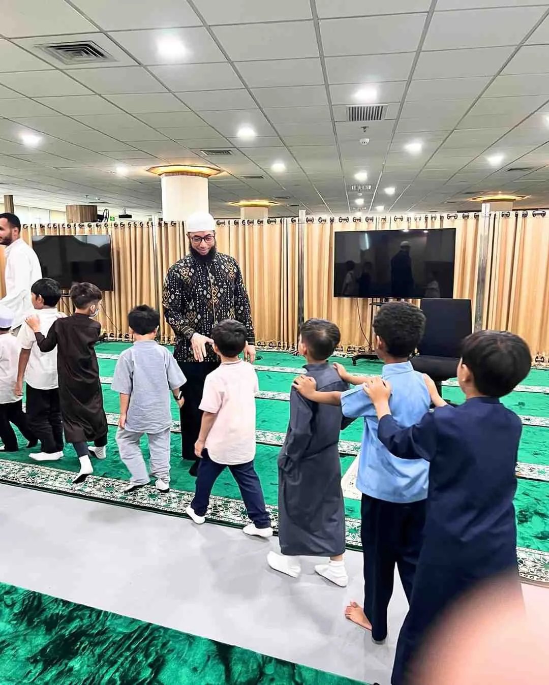A man in a patterned shirt and white cap leading a group of children in a line inside a room with a green carpet, a large television, and beige curtains.