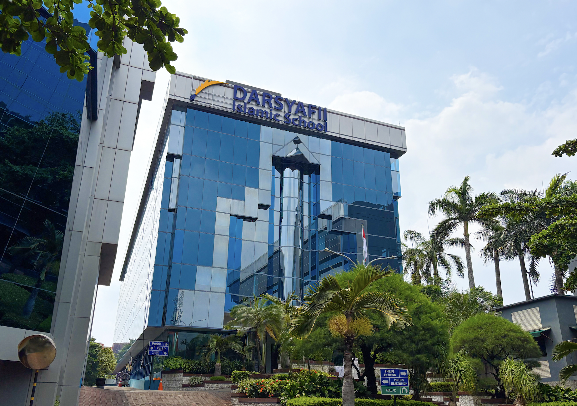 A modern glass building with the sign 'DARYAFI Islamic School' on top, surrounded by lush green trees and a blue sky with some clouds.