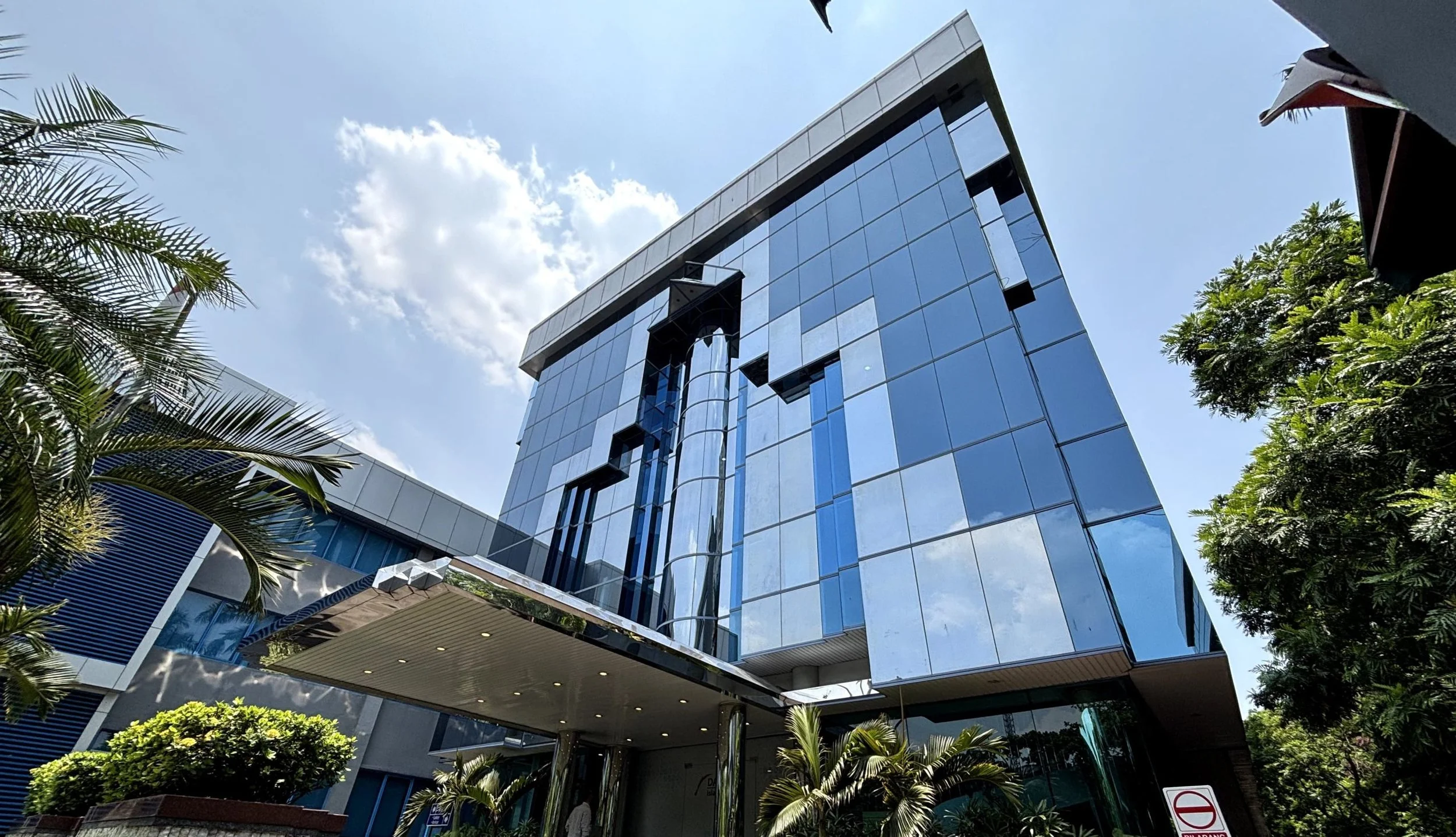 Modern glass office building with a reflective facade, surrounded by lush greenery and palm trees, under a partly cloudy sky.