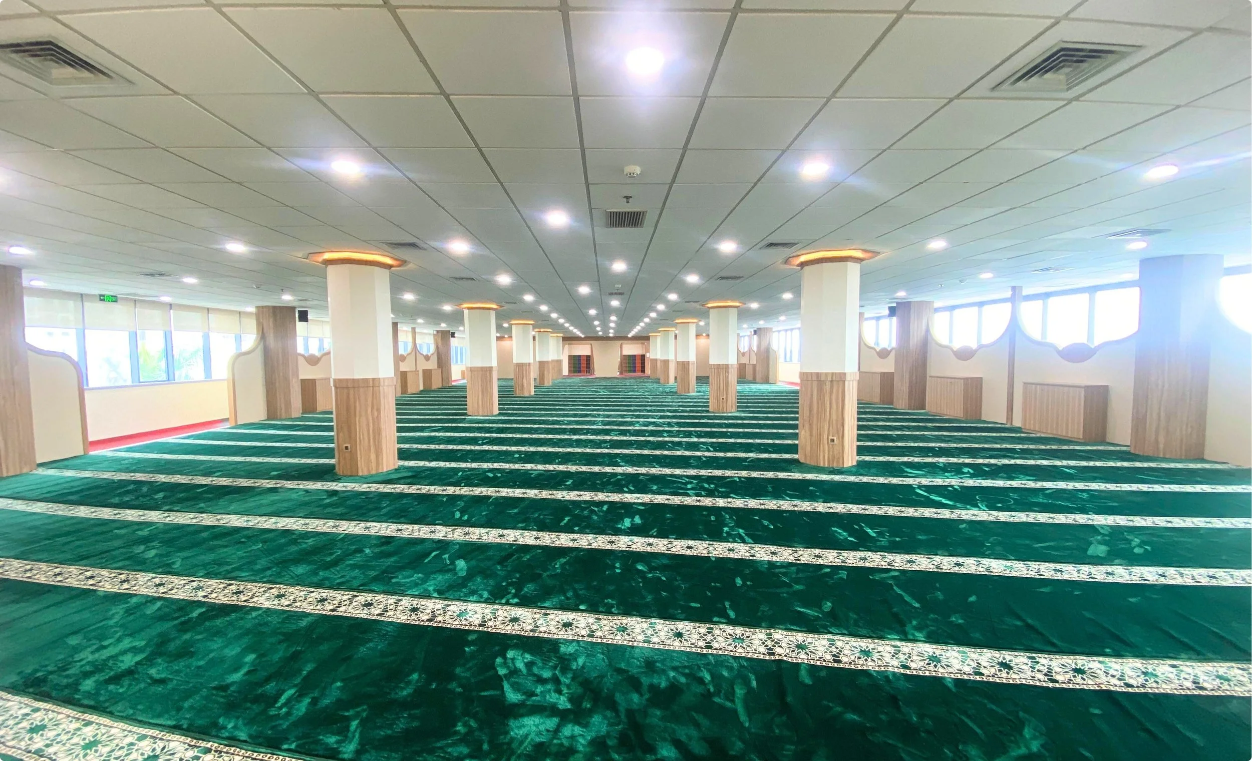 Empty prayer hall or mosque with green patterned carpet, white columns with wood accents, and large windows along the side walls.