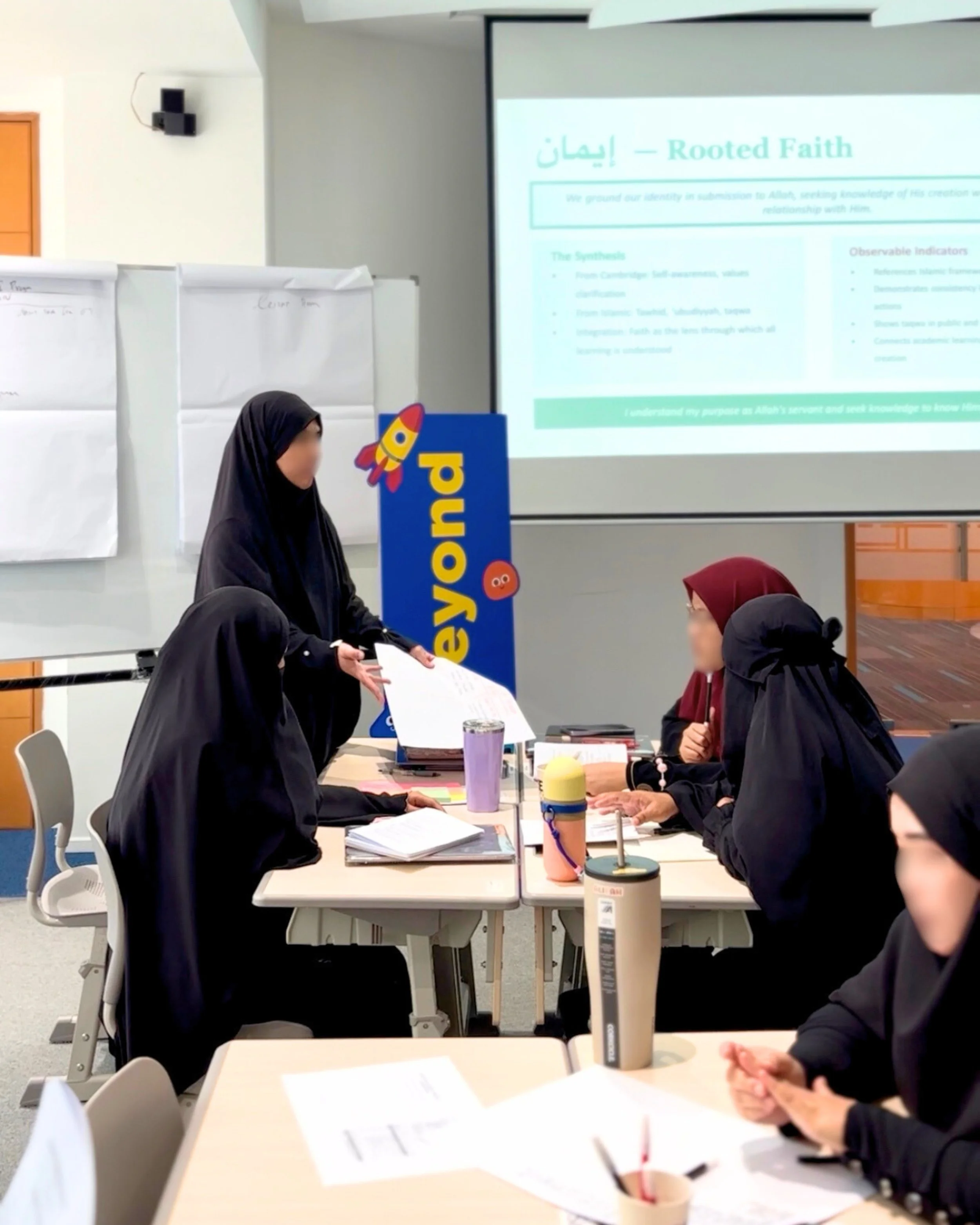 A group of women in hijabs participating in a classroom discussion with a teacher, seated at tables with notebooks and drinks, with a projector screen displaying a presentation about rooted faith.