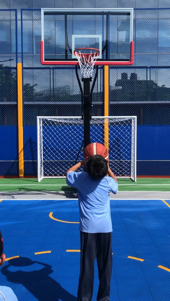 A young boy in a blue shirt prepares to shoot a basketball at a hoop on an outdoor court with blue and green flooring and a small goal attached below the backboard.