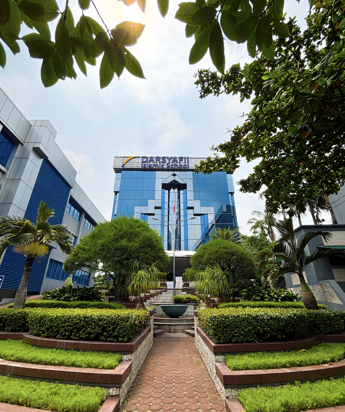 Modern glass building with a sign that reads 'Darsyafi Islamic School,' surrounded by manicured gardens, trees, and a brick walkway leading to the entrance.