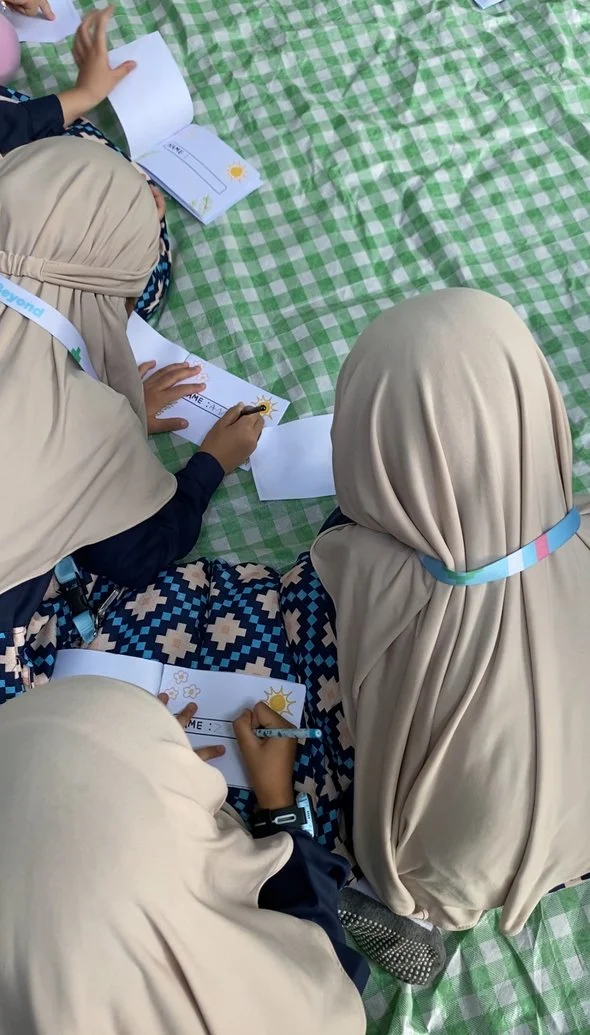 Three children sitting on a green and white checkered blanket, working on white cards with sun illustrations and writing.