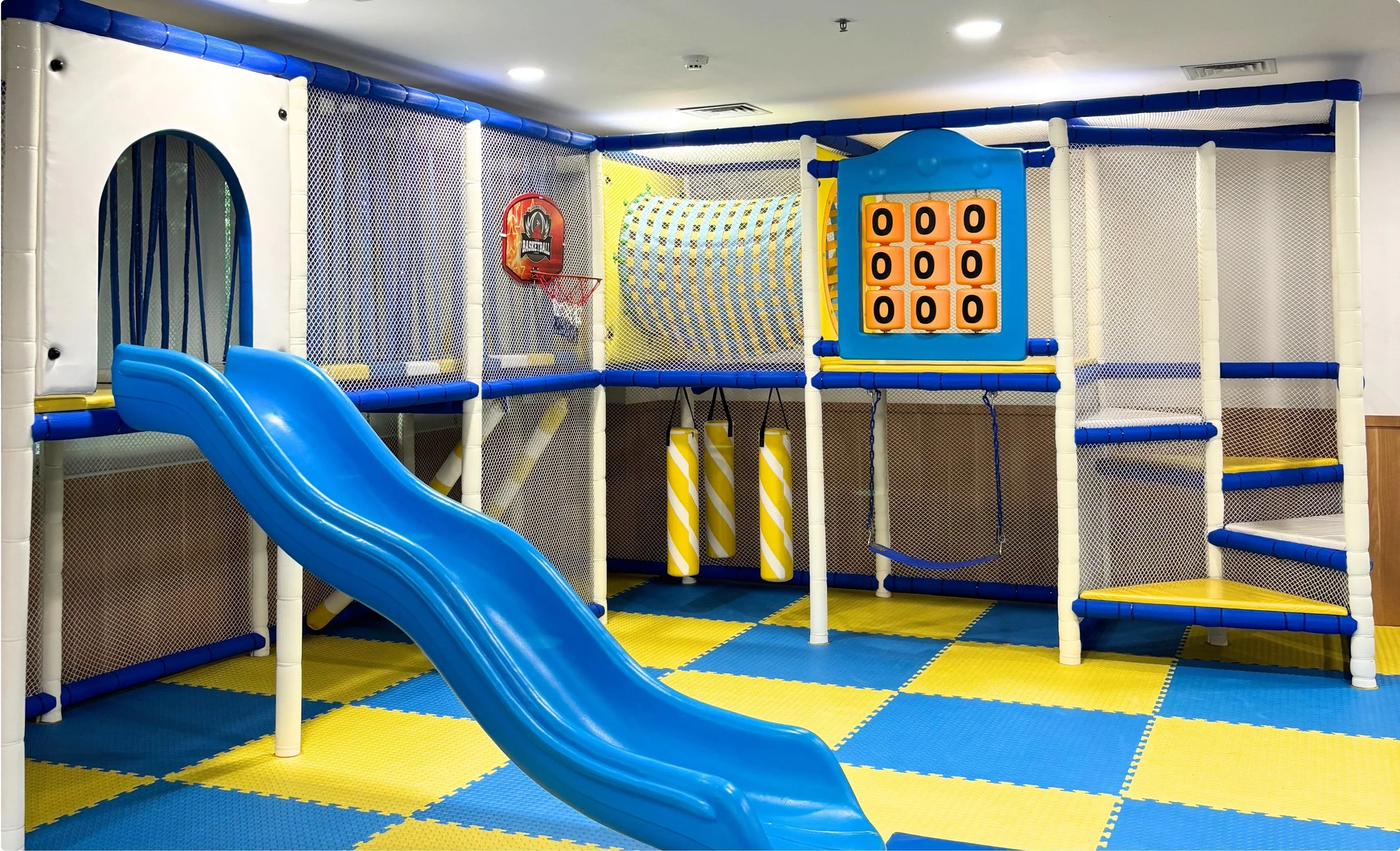 Indoor playground with a blue slide, padded play structure, and climbing areas, enclosed with mesh netting, on yellow and blue checkered flooring.
