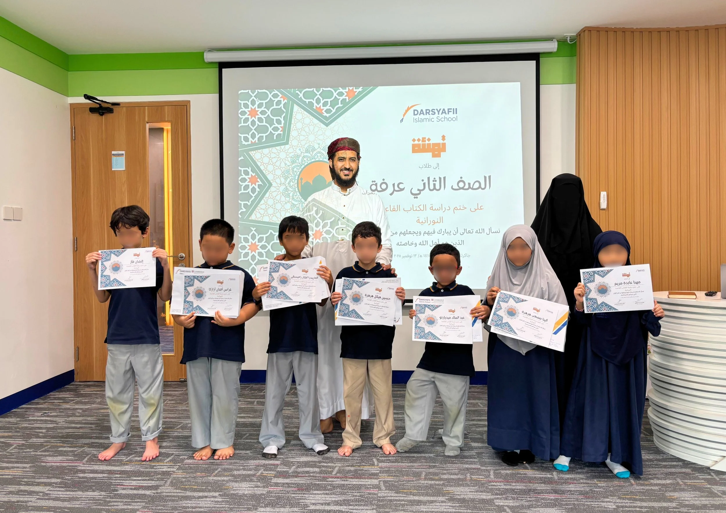 Group of seven children standing in a line holding certificates, with a teacher or adult dressed in black niqab and another woman in gray hijab on the right, in a classroom or auditorium. Behind them is a large screen displaying a presentation with Arabic text, a photograph of a man, and the logo of Darsyafi Islamic School.