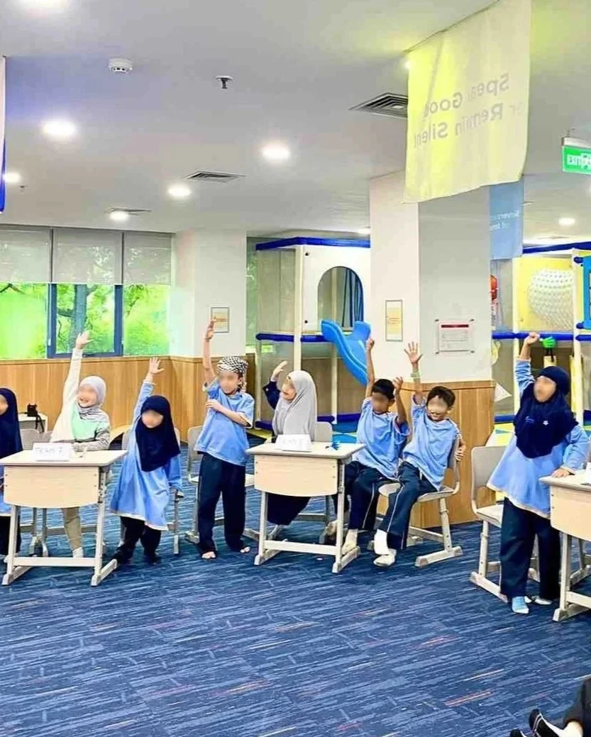 Children sitting and standing at desks in a classroom, raising their hands, with a play area and windows in the background.