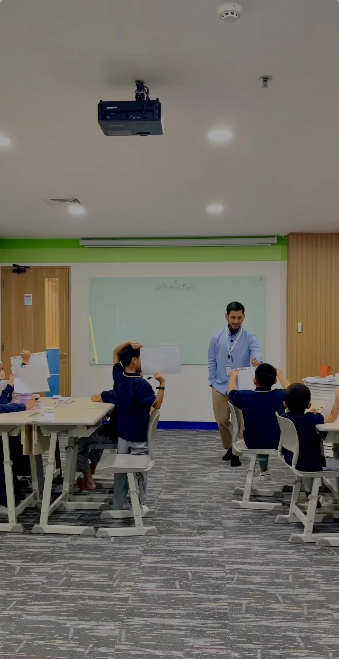 Classroom scene with students seated at desks, teacher standing and engaging with students, whiteboard at the front, ceiling projector, and cabinets on the side.