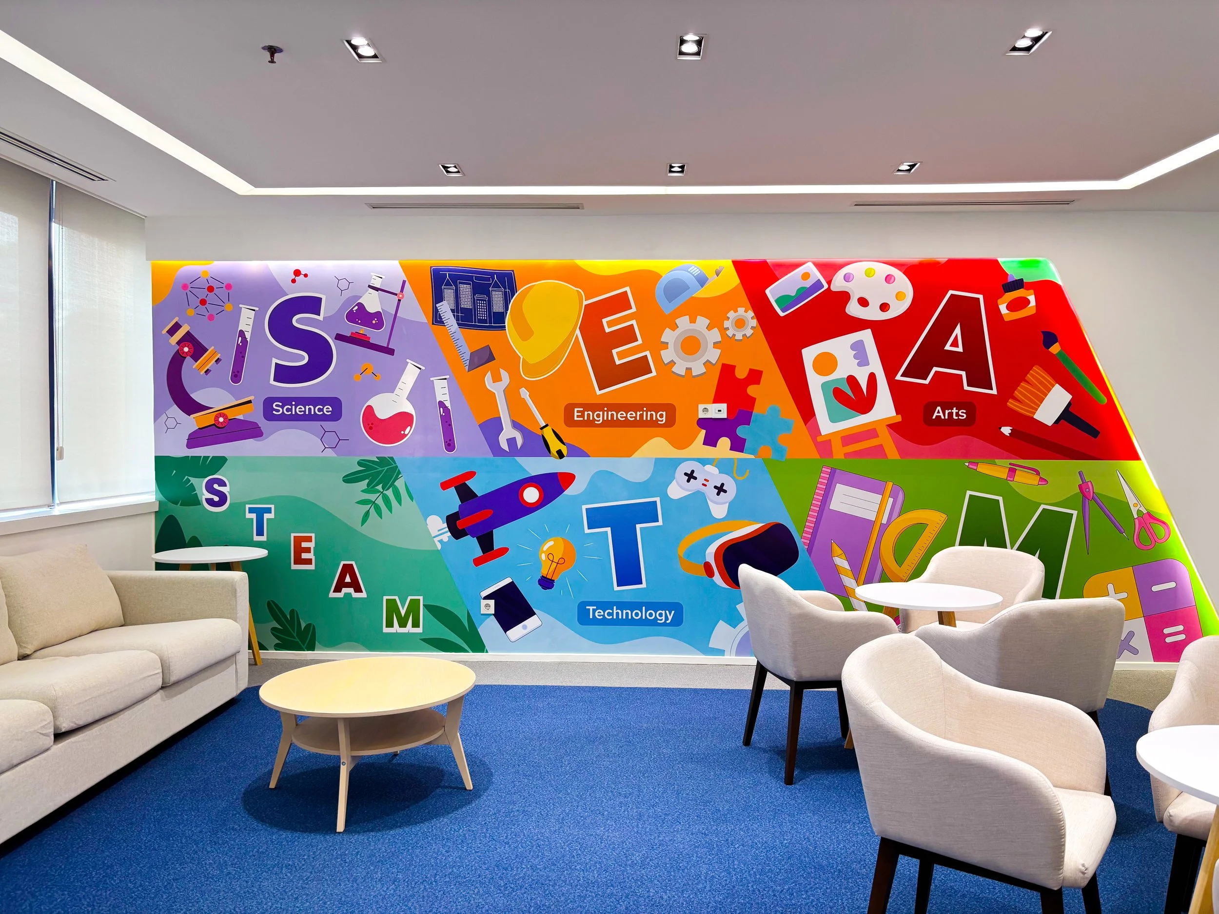 Colorful mural on a wall with words and icons representing science, engineering, arts, and technology in a modern office or waiting area. The mural includes symbols like a microscope, rocket, paint palette, gears, and a light bulb. There are couches, chairs, and tables in the room.