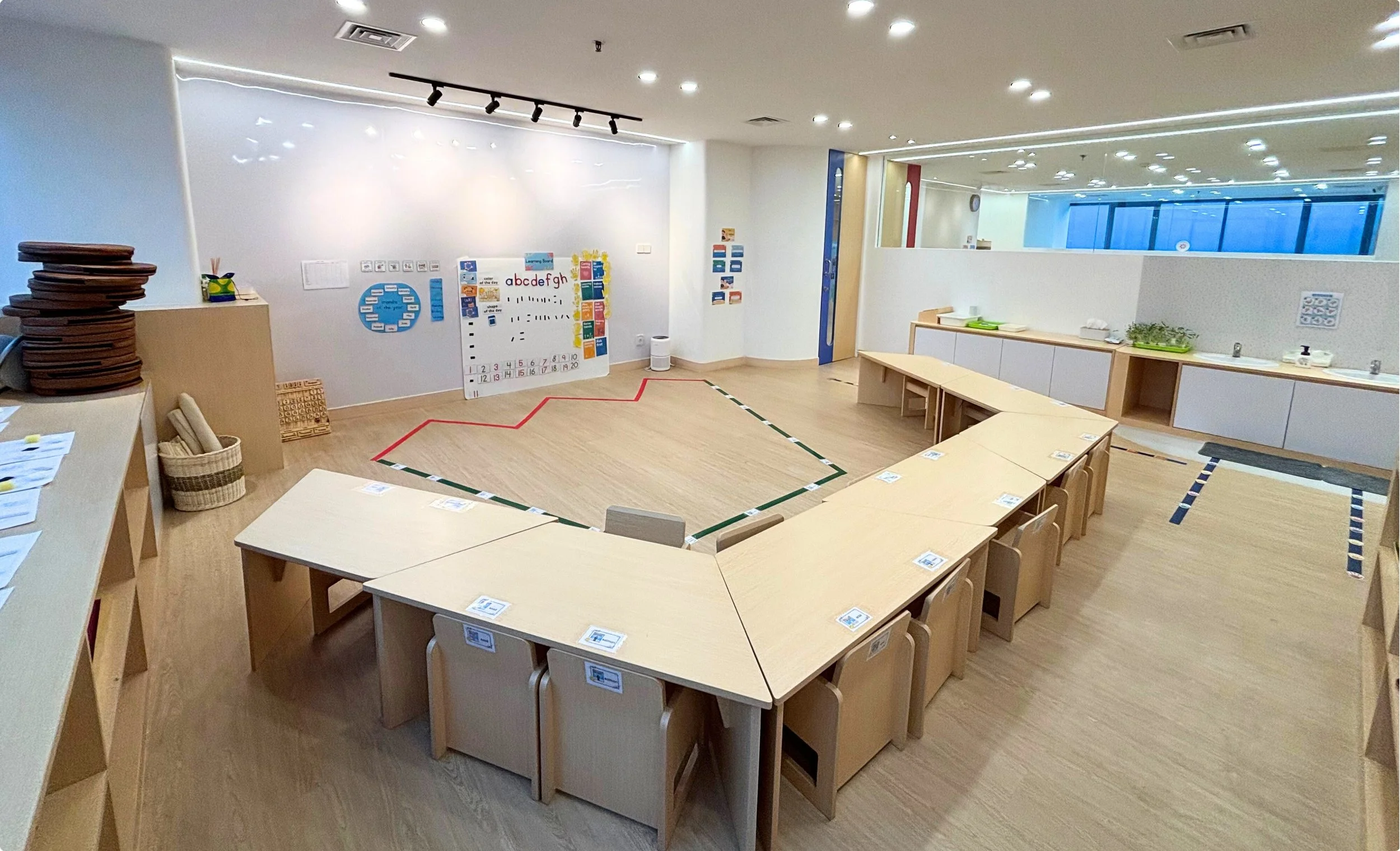An empty classroom with wooden tables arranged in a U-shape, small chairs, educational posters on the wall, and a red line marking on the floor.