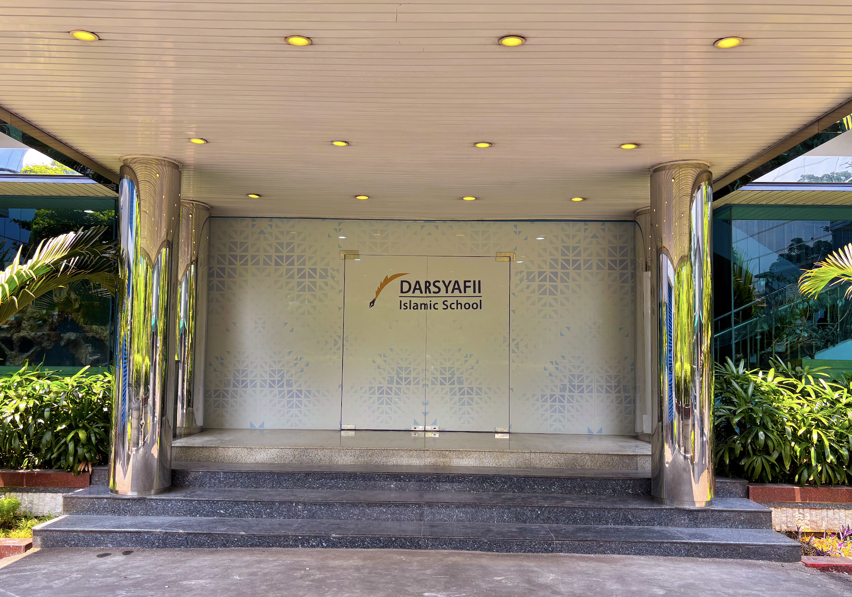 Entrance to Darsyafii Islamic School with glass doors, plants on sides, steps leading up, and ceiling lights.