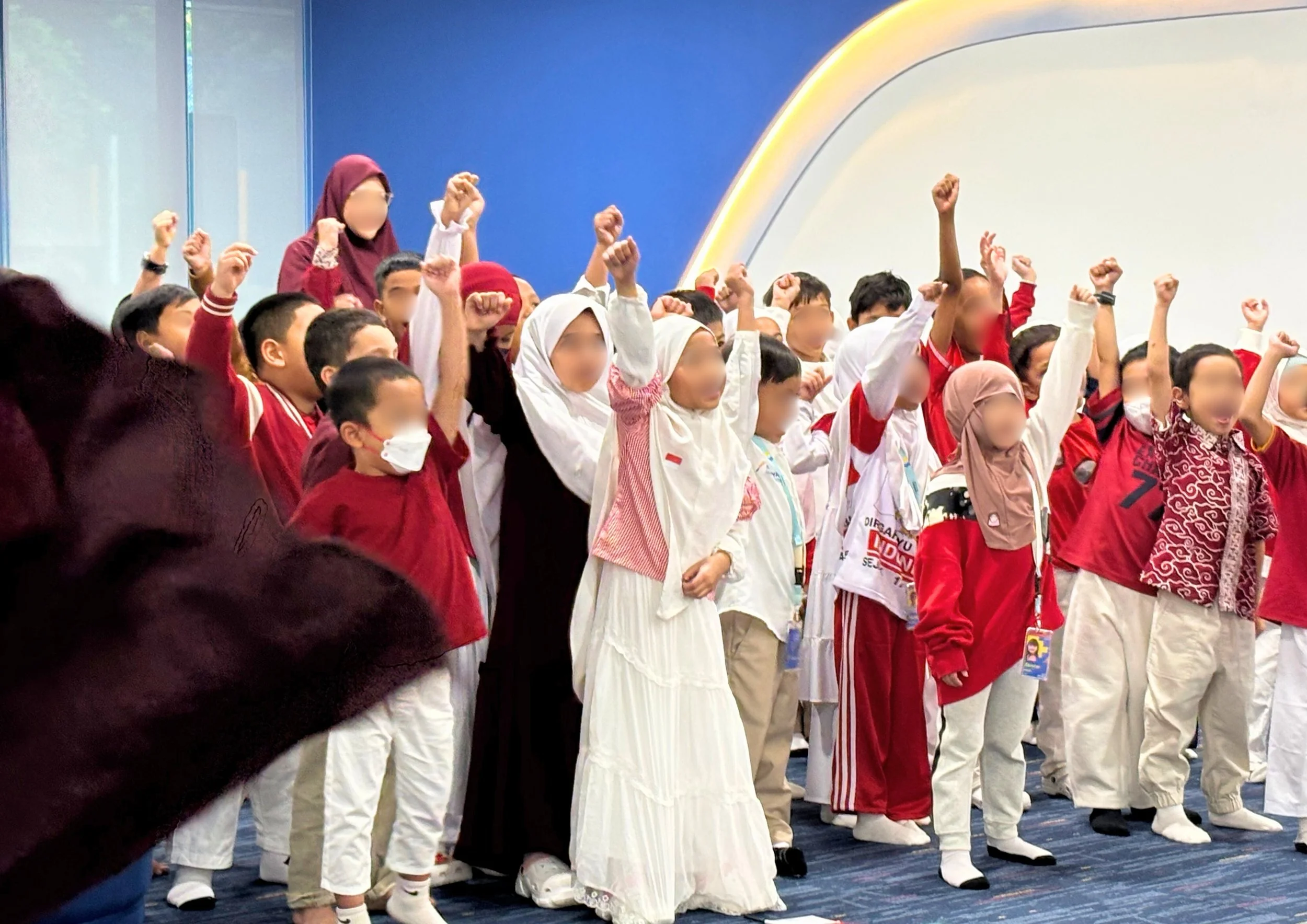 Group of children dressed in red and white, some wearing masks or headscarves, standing on a stage with their arms raised in victory or celebration.