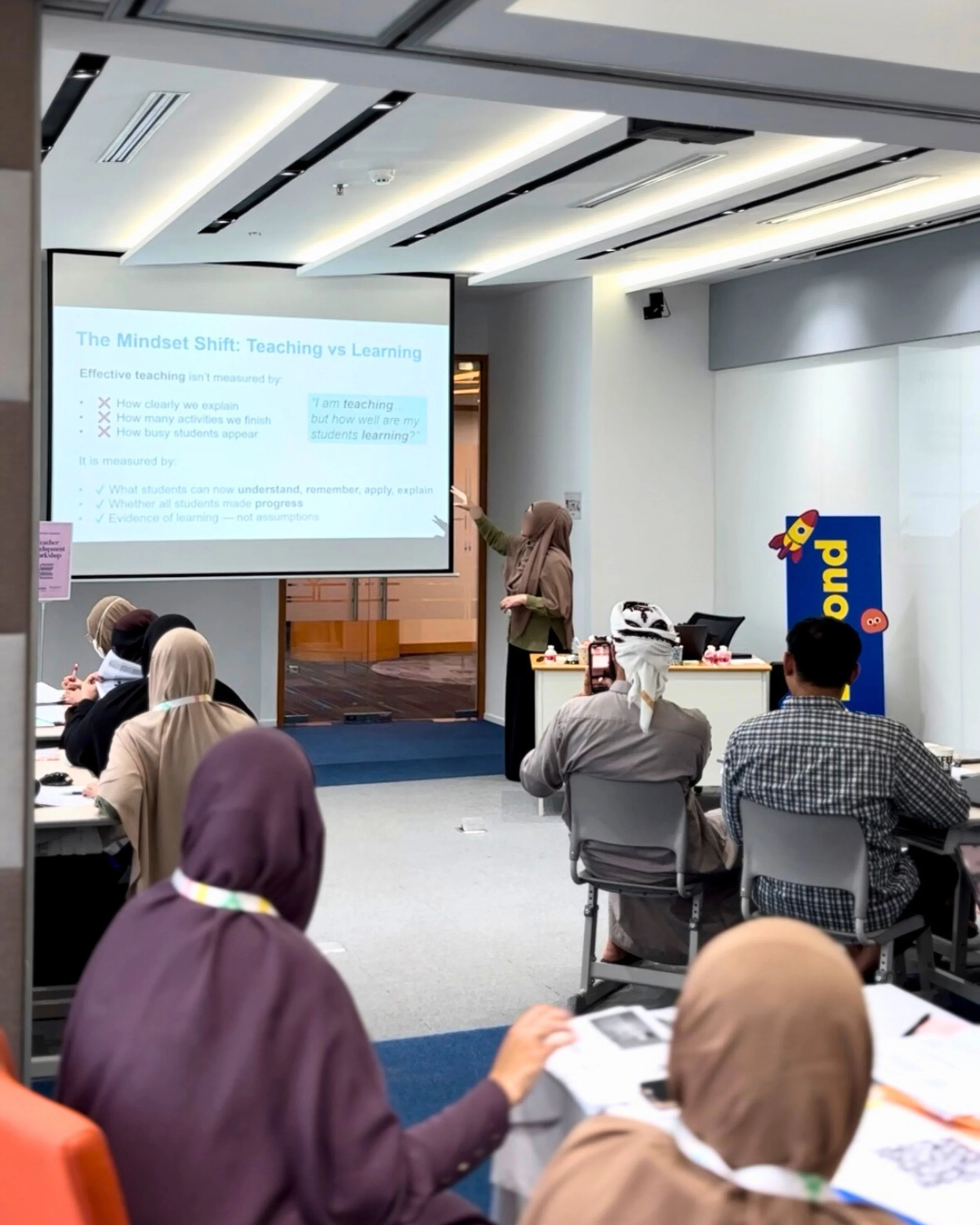 A woman giving a presentation on stage, pointing at a slide titled 'The Mindset Shift: Teaching vs Learning' in a conference room, with several seated attendees, some taking notes and one recording with a phone.